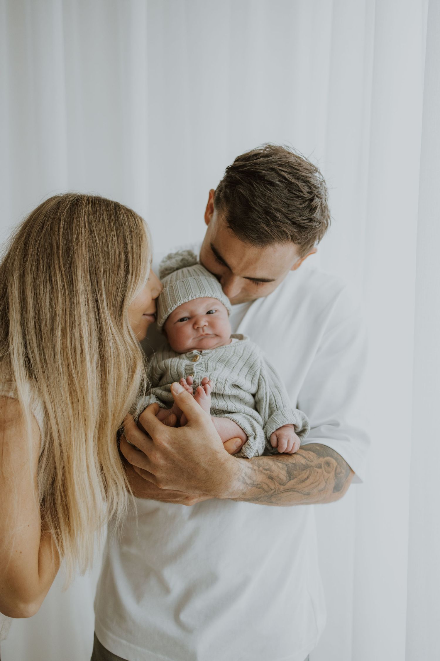 Parents tenderly holding newborn baby against white curtain background.