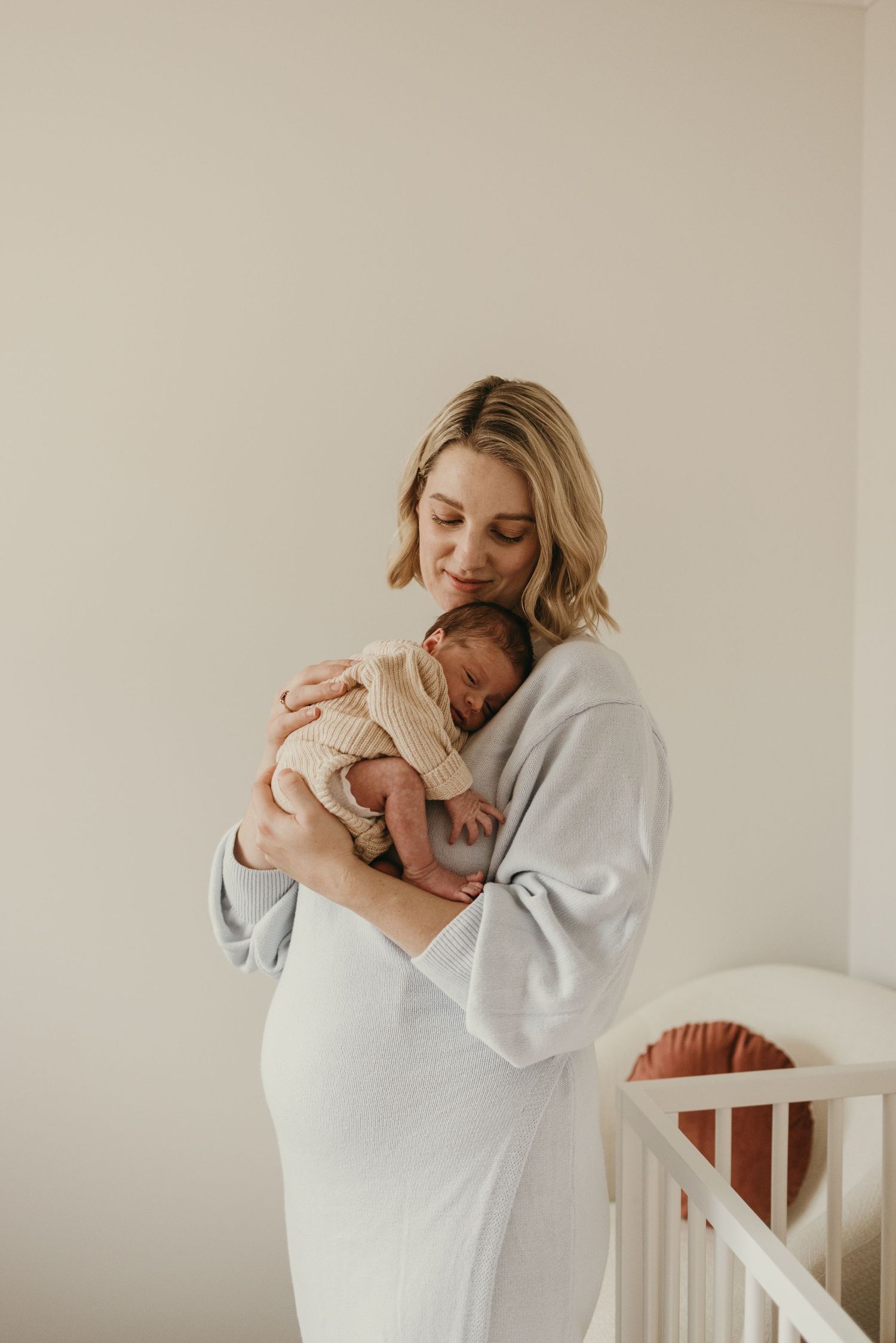Parent wearing white embraces newborn baby next to a white crib in a softly lit nursery.