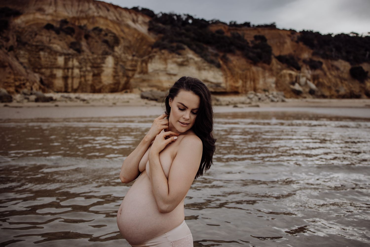 Pregnant woman cradling her bump on a beach with rocky cliffs during a coastal maternity photoshoot.