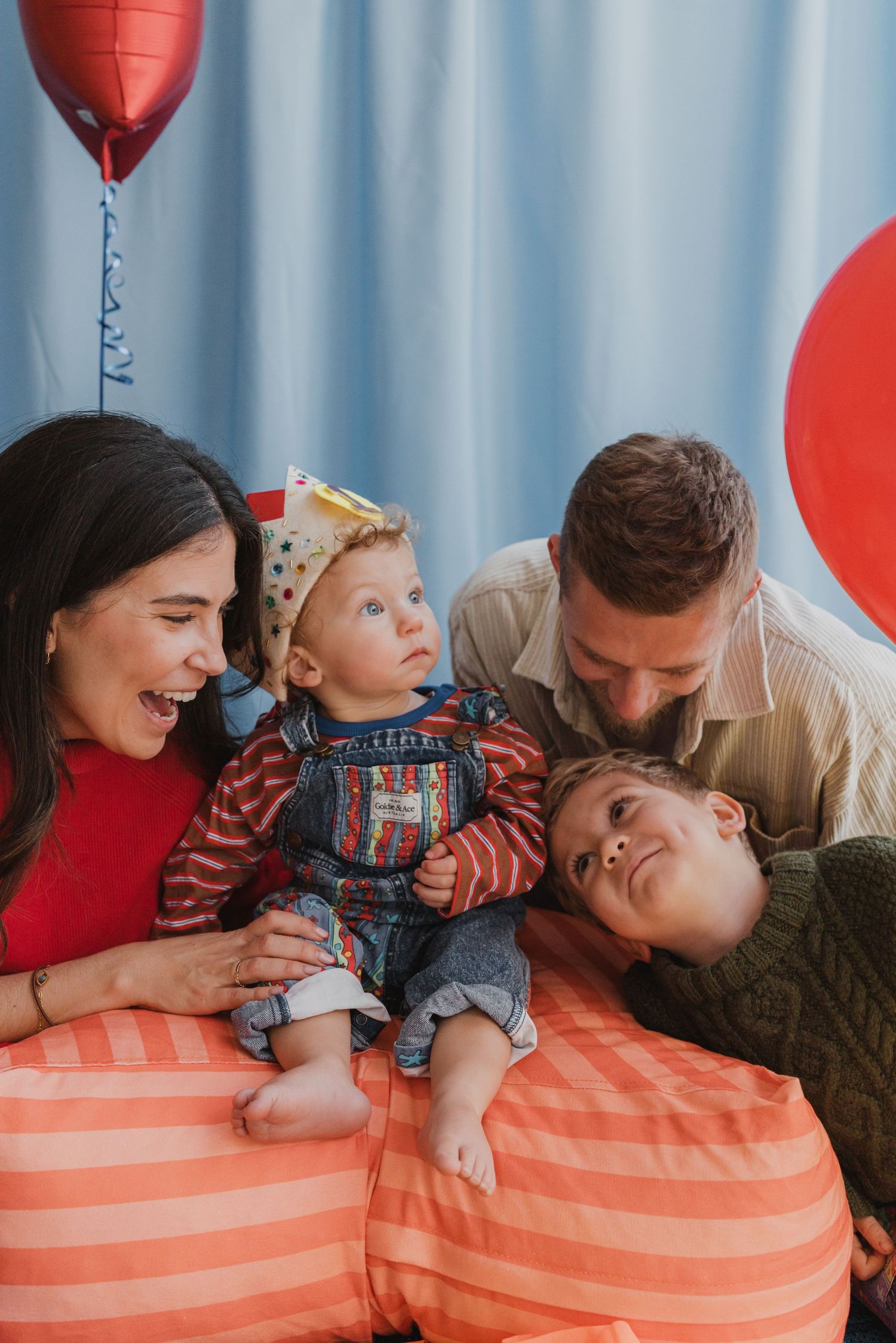 A joyful family celebrating a baby's first birthday with red balloons and a party hat.