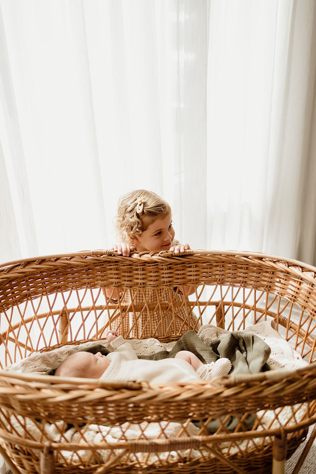 A toddler girl in a rattan bassinet peers down at a sleeping newborn baby wrapped in soft blankets.