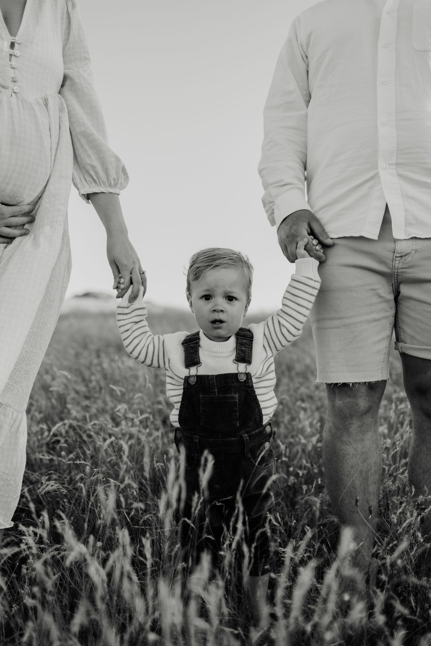 Black and white photo of a toddler in overalls holding parents hands in a field.