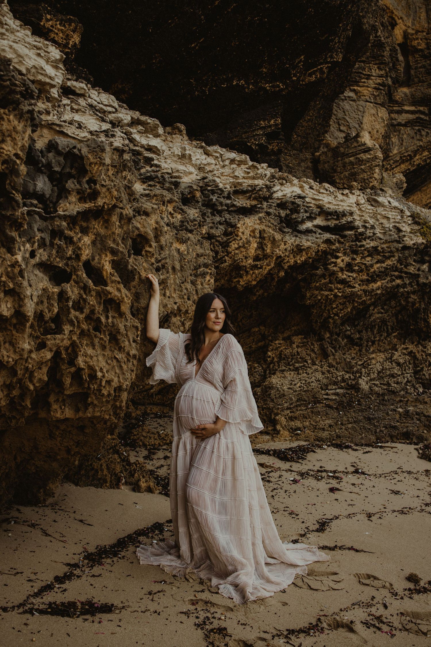 Pregnant woman in white boho dress poses gracefully against rocky cave walls on sandy beach.