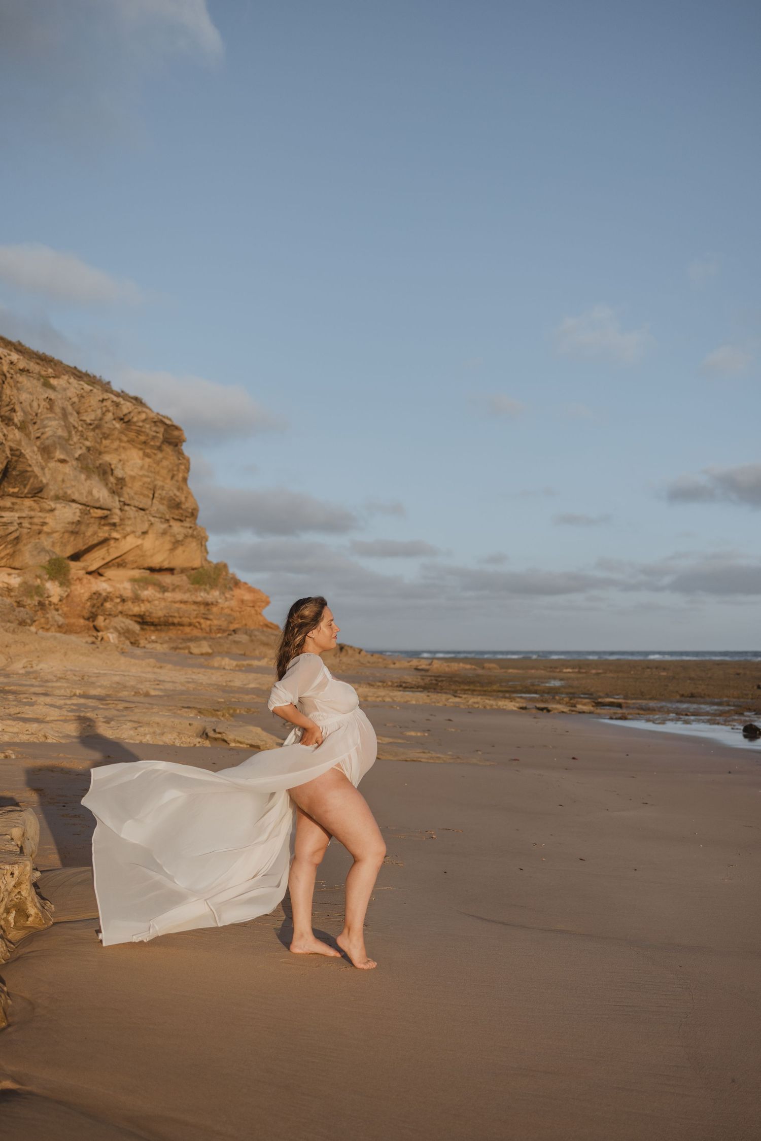 Pregnant woman in flowing white dress on sandy beach with rocky cliffs at golden hour.