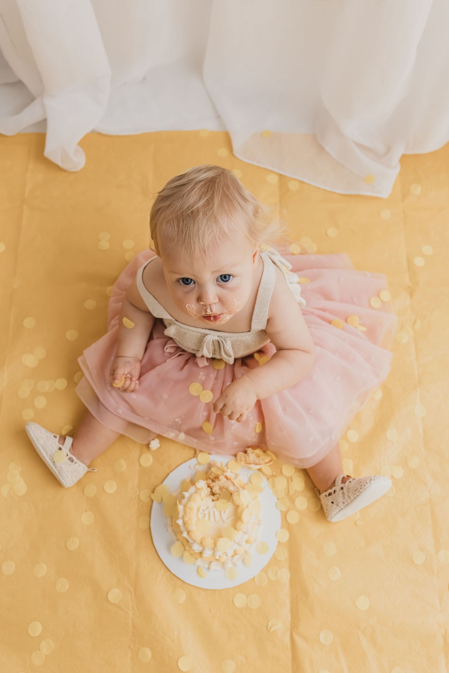 Baby girl in pink tutu dress sitting on yellow blanket with smash cake for first birthday photo.
