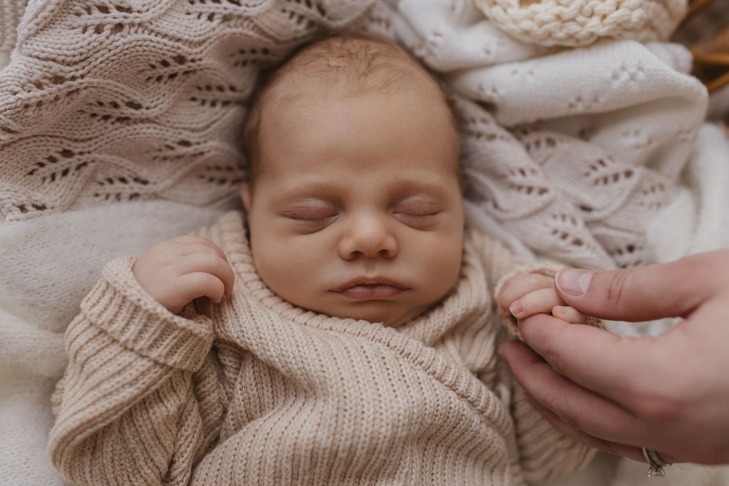 Peaceful newborn baby sleeping in a beige knit wrap on a soft blanket, gently holding an adult's finger.