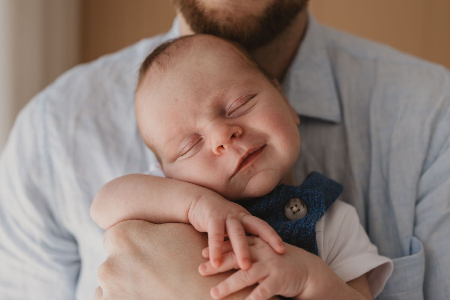 A sleeping newborn baby smiles peacefully while being cradled in a bearded fathers arms, wearing a denim overalls outfit.