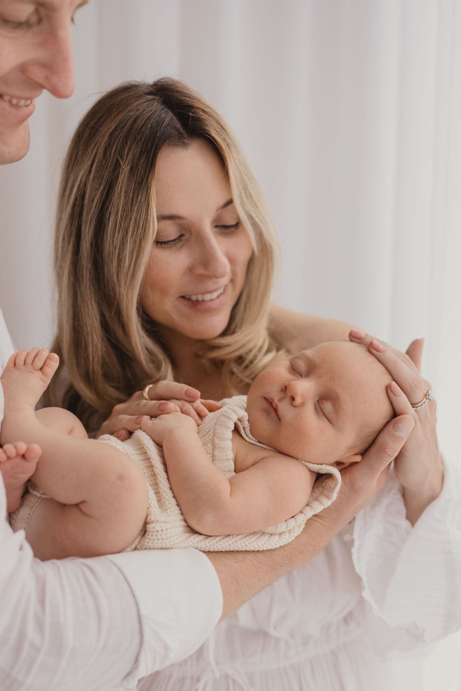 Parents tenderly hold their sleeping newborn baby in white clothing during a professional family portrait session.