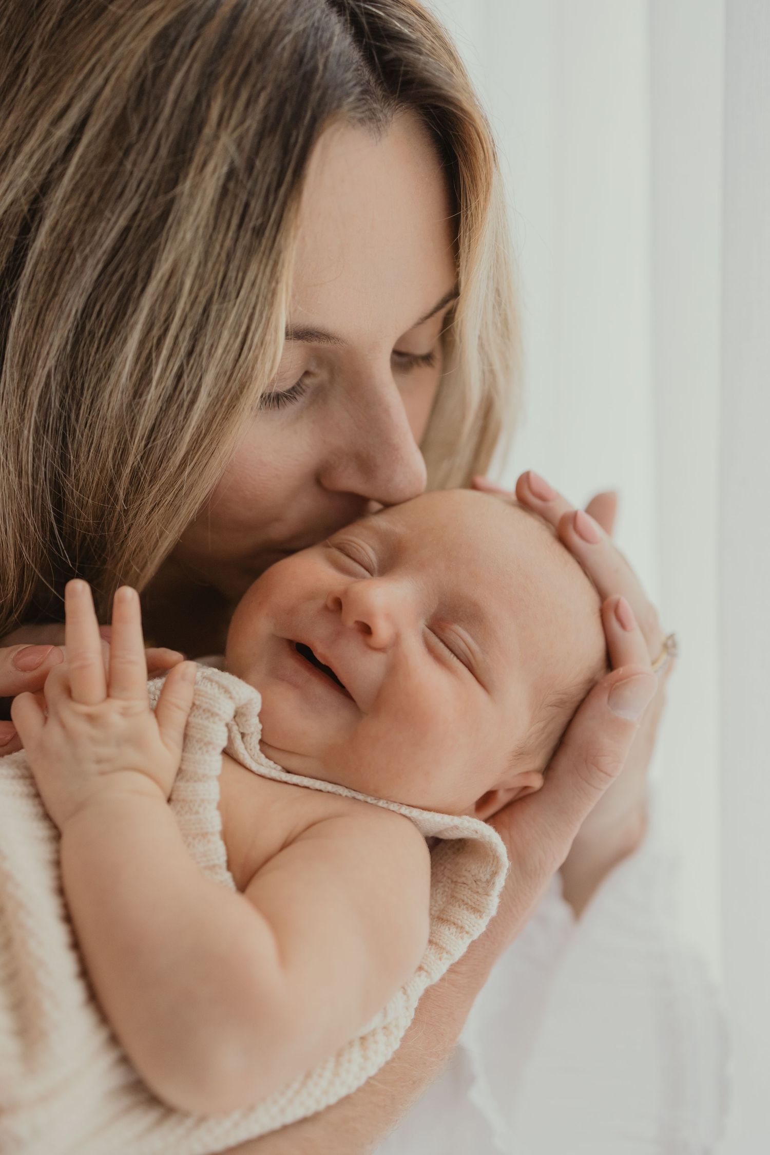A mother tenderly kisses her smiling newborn baby, cradling the infant gently in her hands.