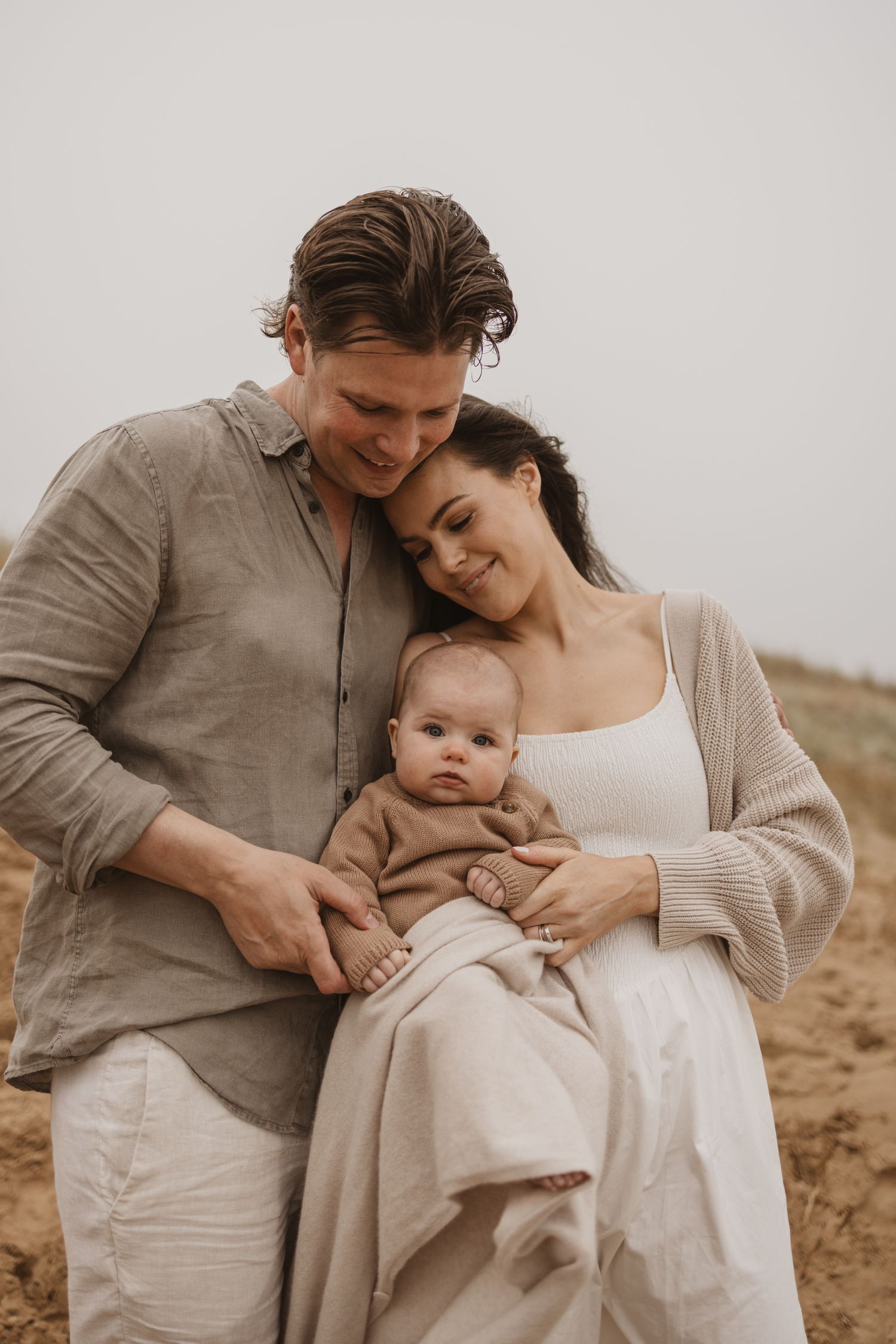 A young family in neutral tones tenderly holds their newborn baby outdoors on a sandy beach.