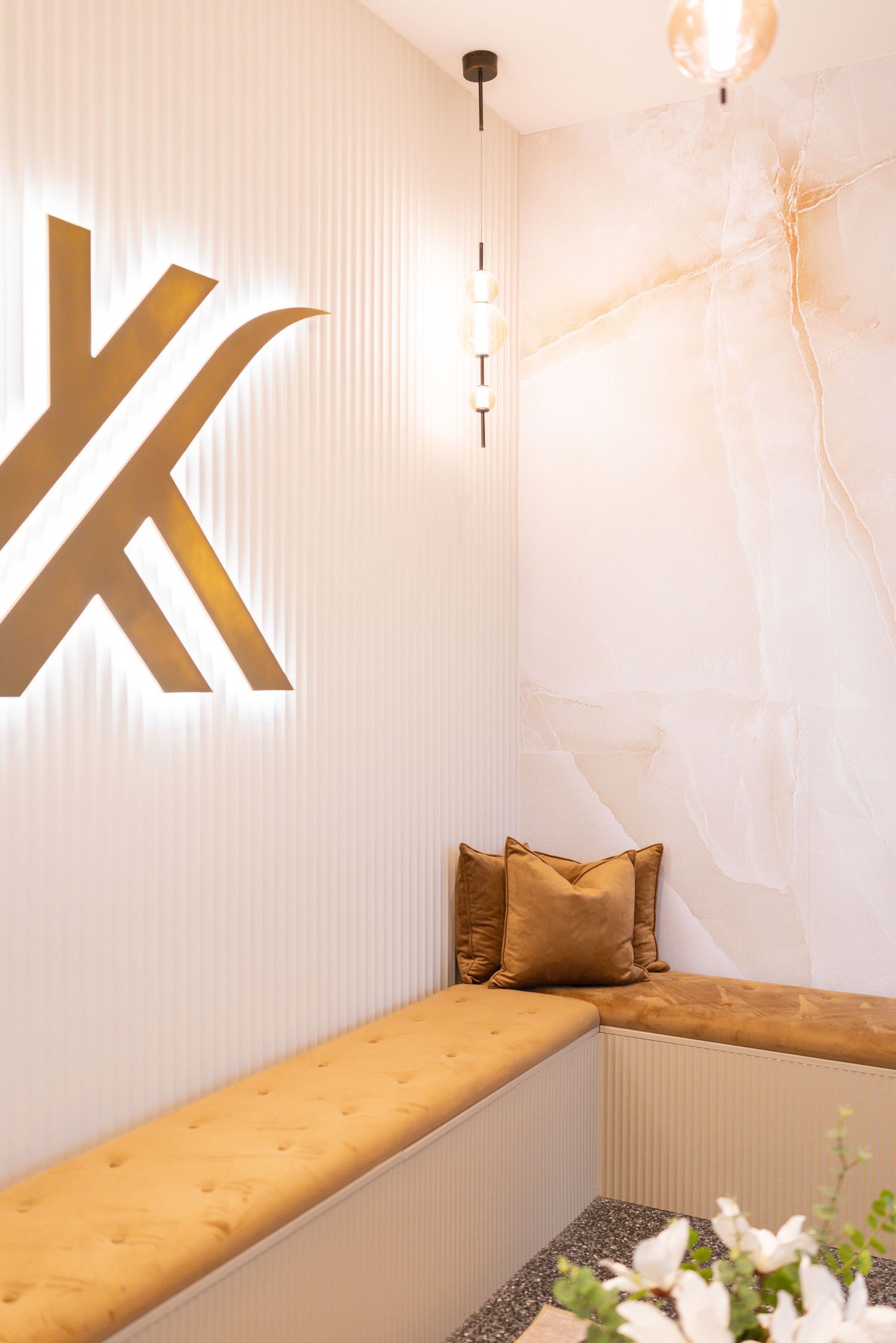 Elegant salon waiting area with gold logo, tufted bench, velvet cushions, marble wall, and pendant lighting.