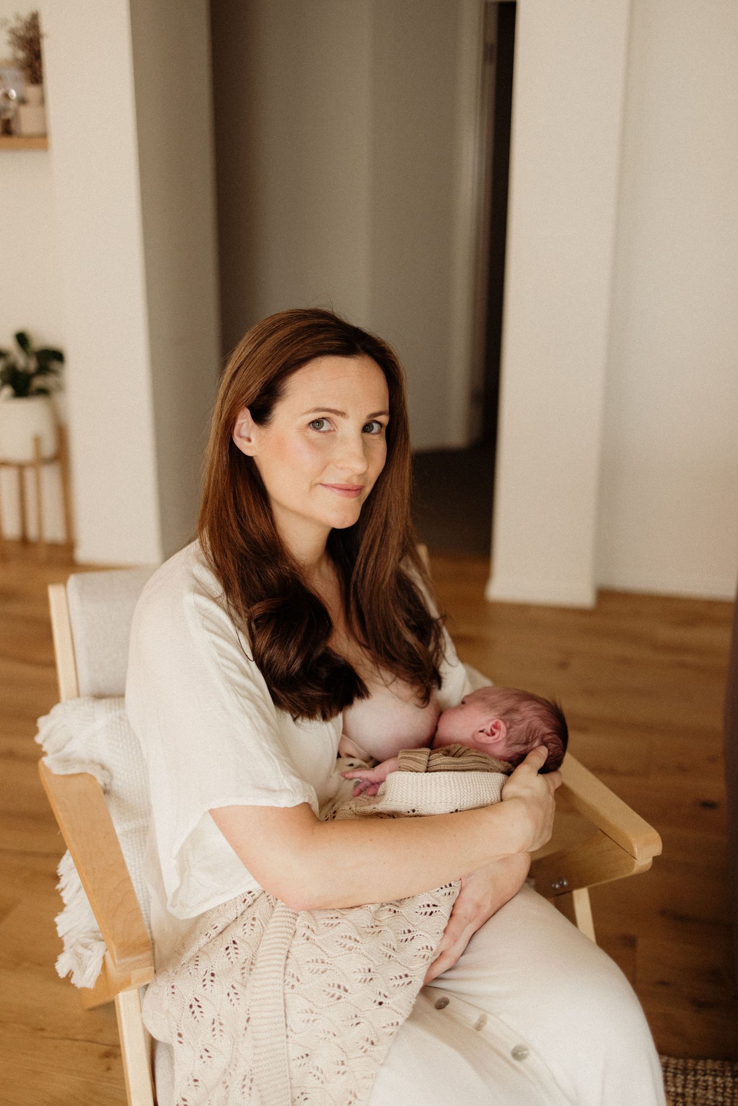A mother in a white dress sits in a wooden chair breastfeeding her newborn baby in a bright modern home.