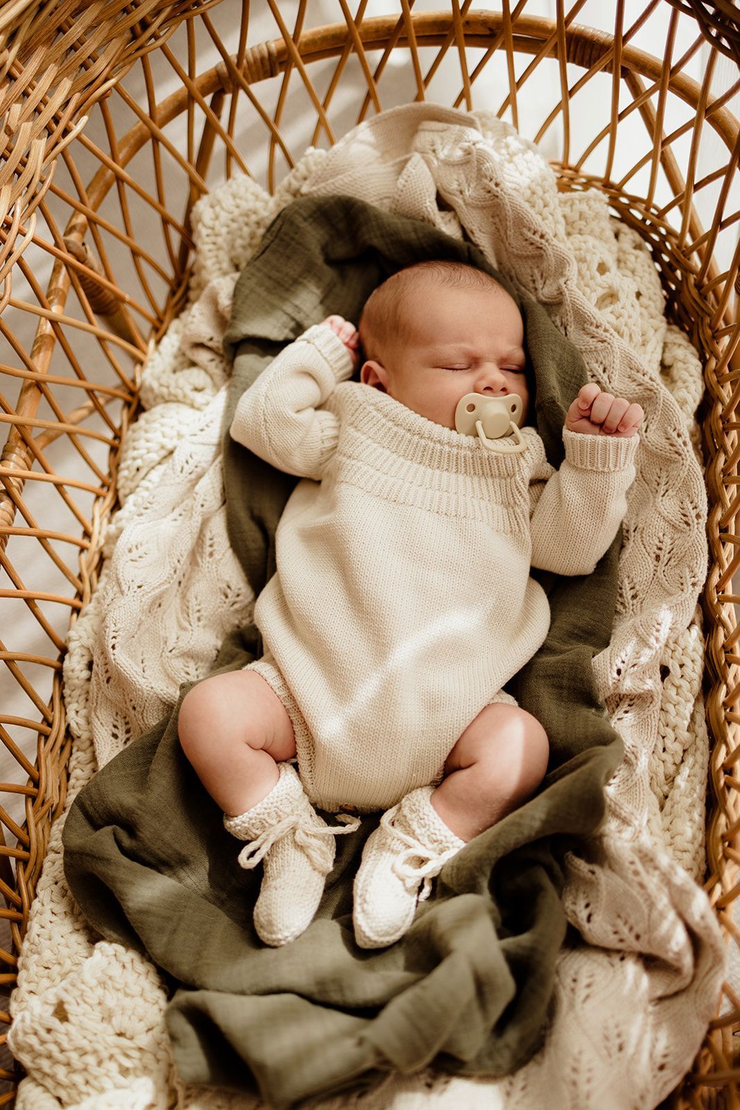 Newborn baby sleeping peacefully in a wicker bassinet, dressed in a cream knit outfit with a pacifier.