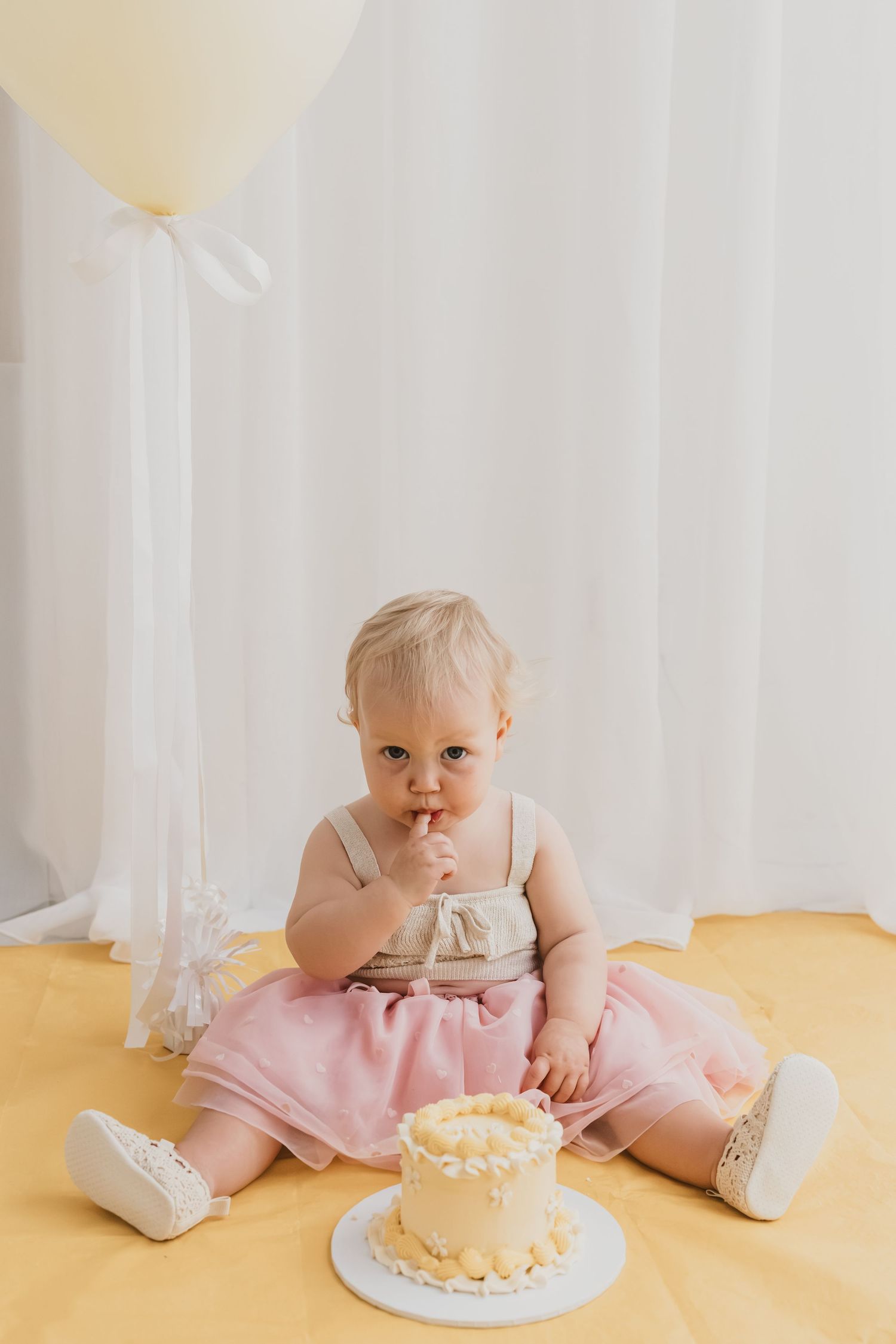 Baby girl in pink tutu sits on yellow floor tasting her first birthday cake near a white balloon.
