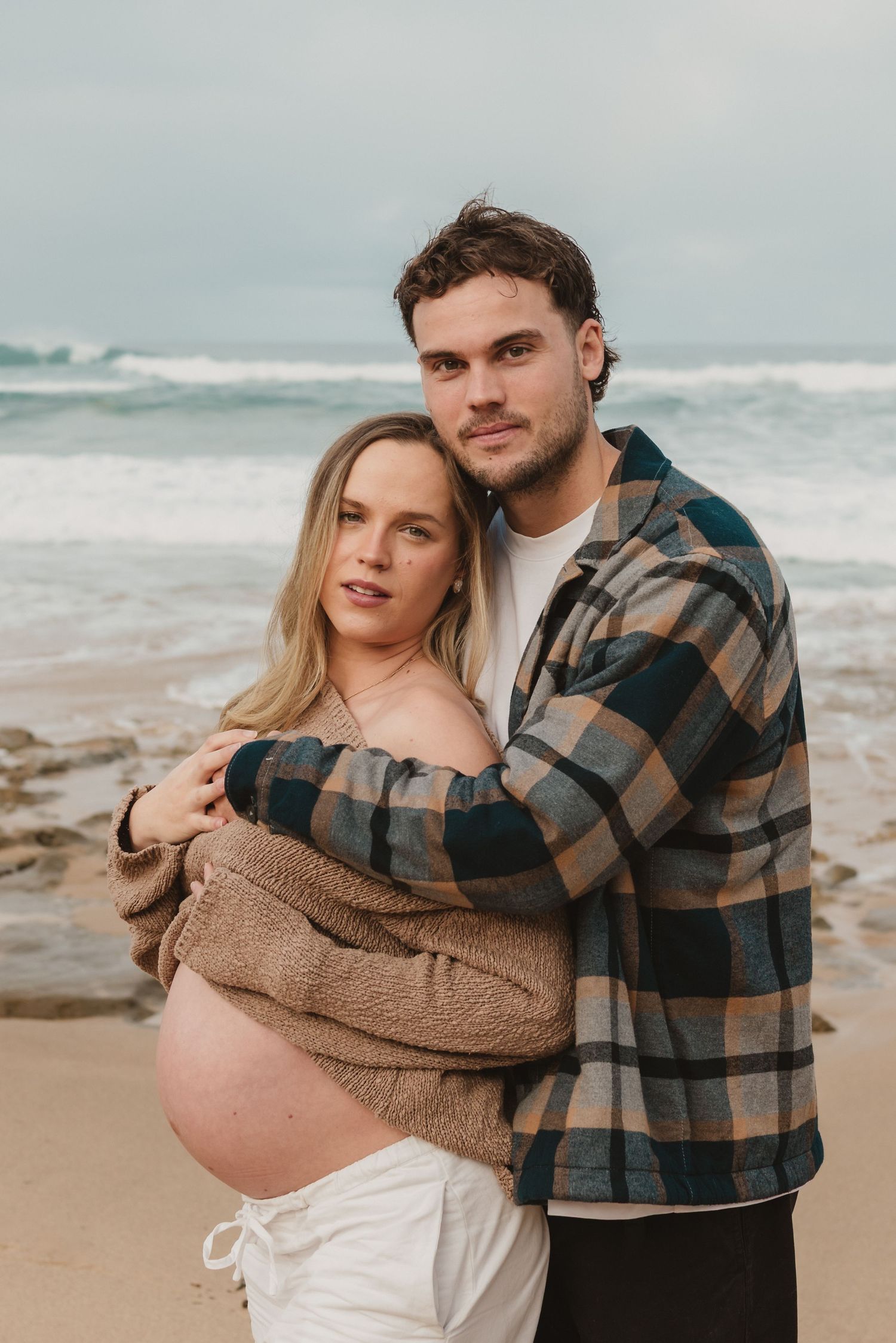 Pregnant couple embracing on a beach, man in plaid shirt hugging woman in tan sweater with ocean waves behind them.