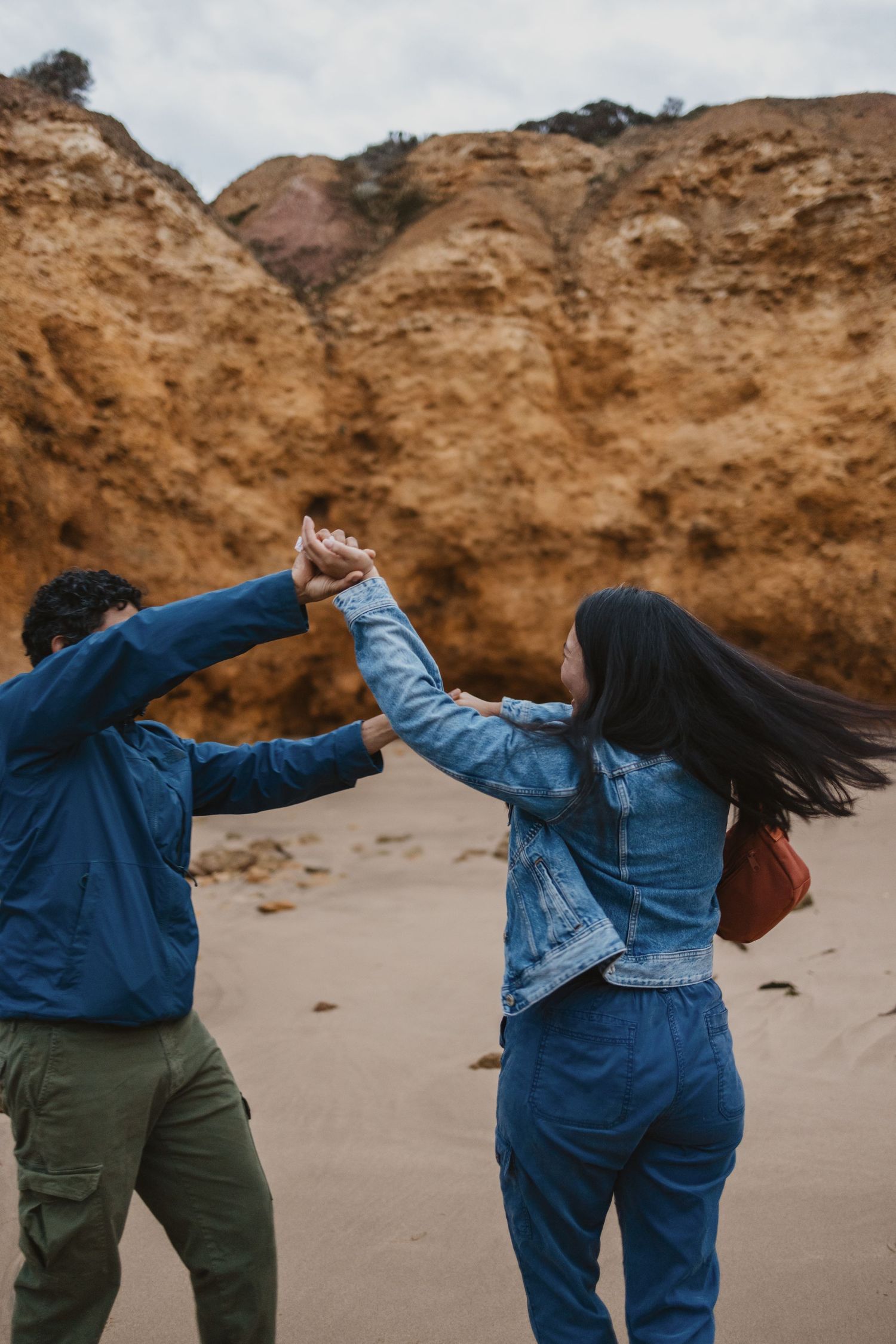 Couple dancing on sandy beach with dramatic orange cliffs backdrop, both wearing blue denim jackets, holding hands up.