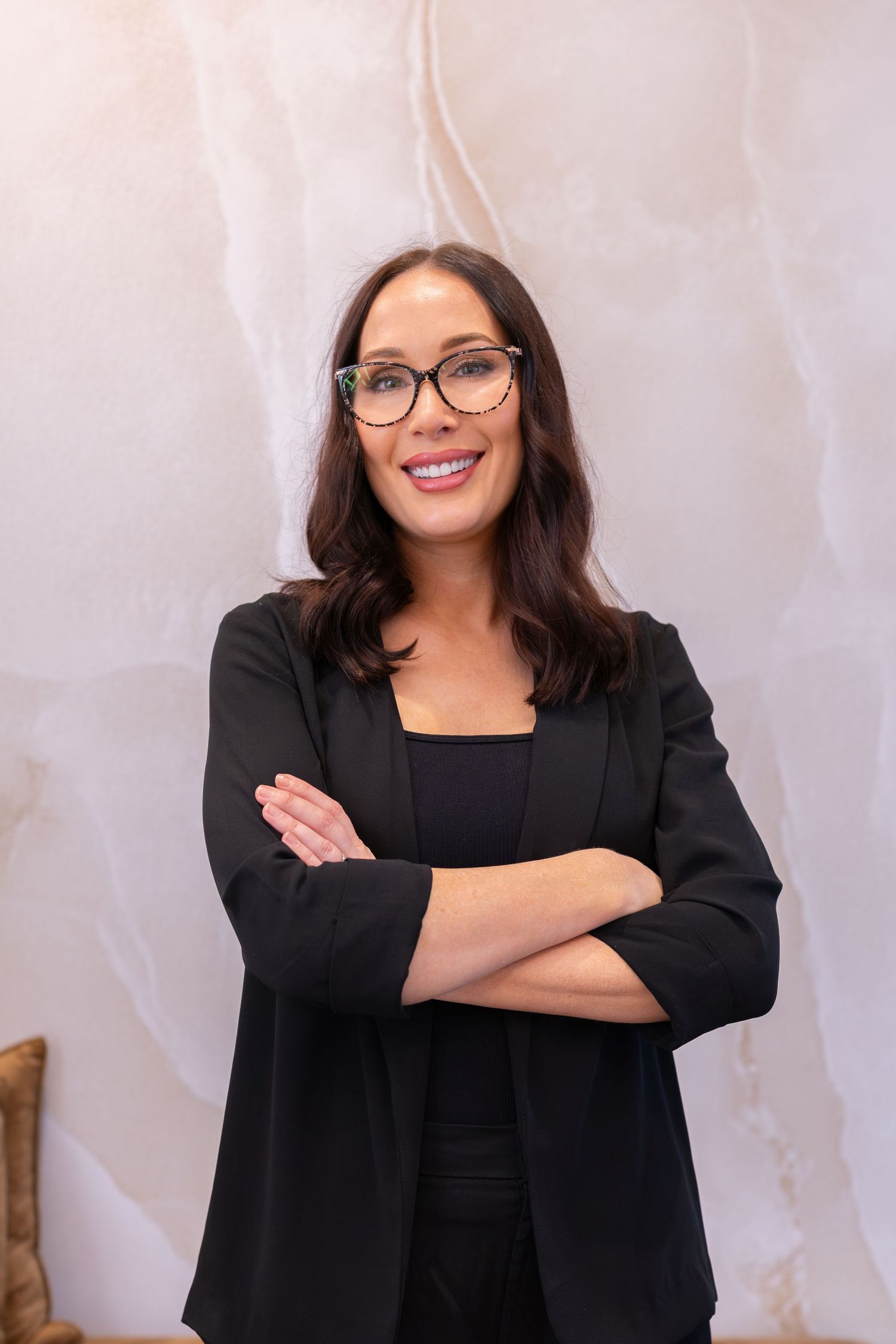 Professional woman in black blazer and glasses smiling with arms crossed against marble background.