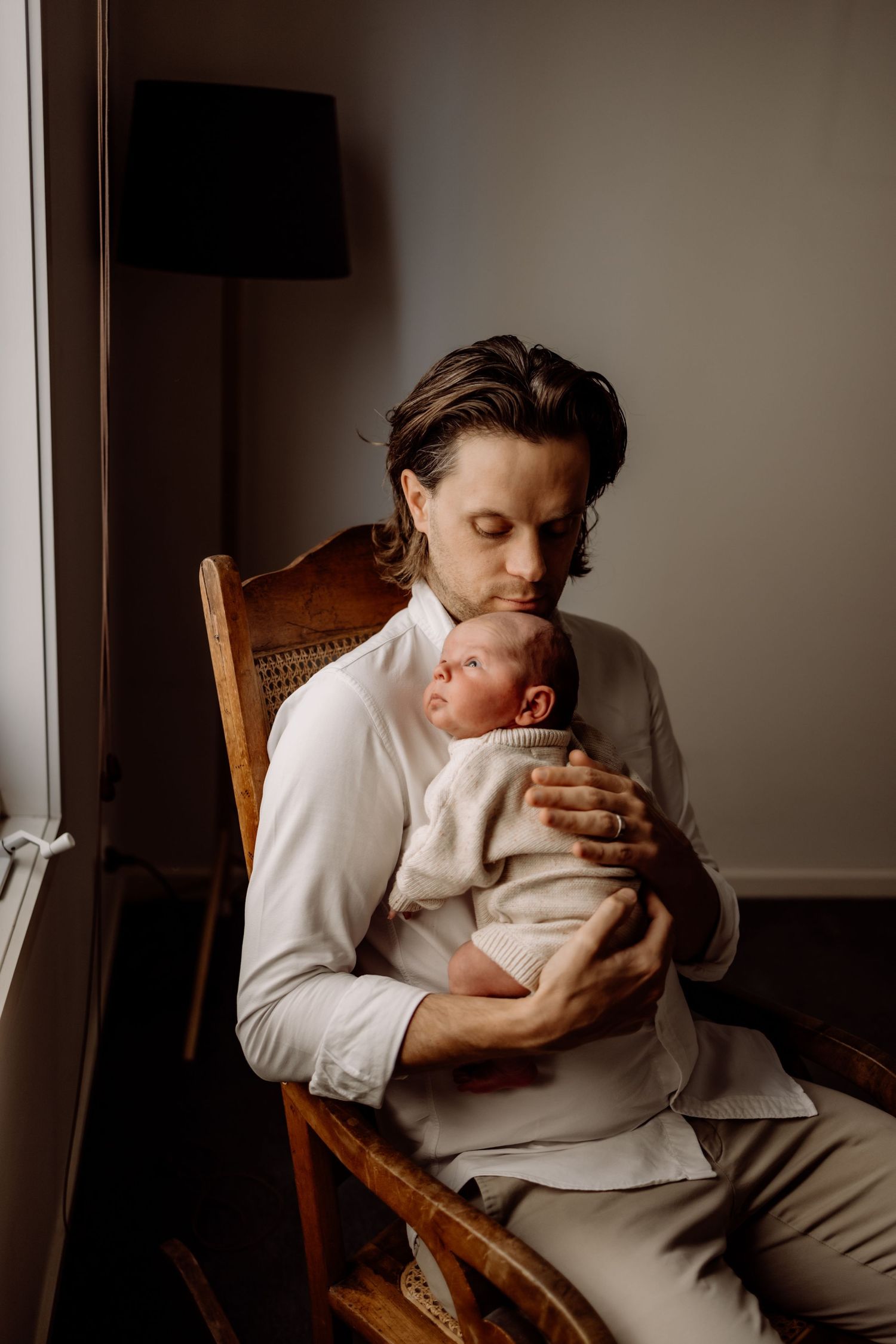 A father gently kisses his newborn baby while sitting in a wooden rocking chair in a softly lit room.