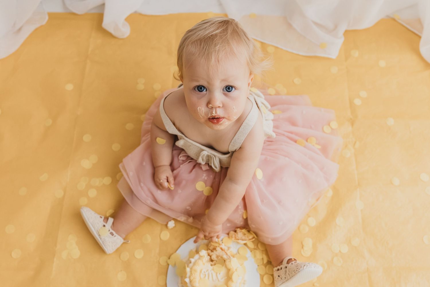 Adorable baby girl in pink tutu dress looking up after smashing her first birthday cake on yellow backdrop.