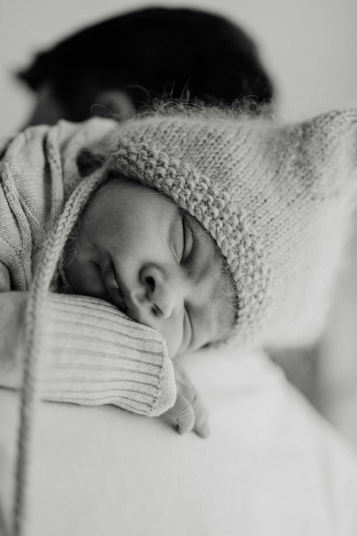 Black and white photo of a sleeping newborn baby wearing a knit bonnet, cradled in a parent's arms.