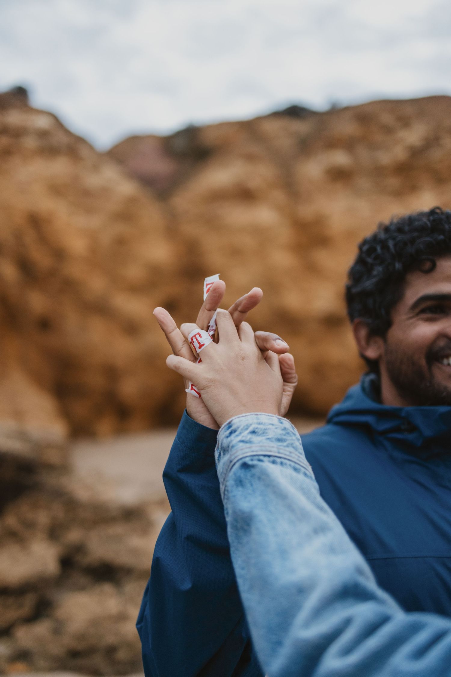 Couple interlocking fingers outdoors near rocky terrain, man in blue jacket smiling, woman with red nail polish.