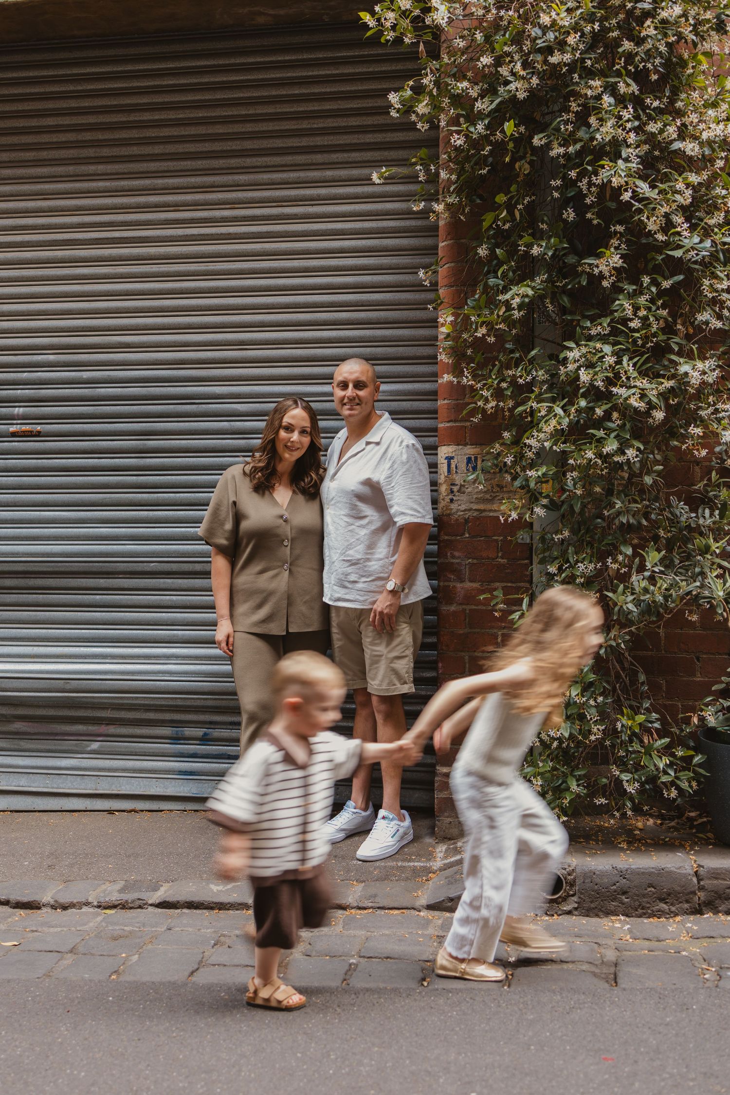 Family portrait with two adults smiling and two children playing on a sunny urban street near a brick wall.