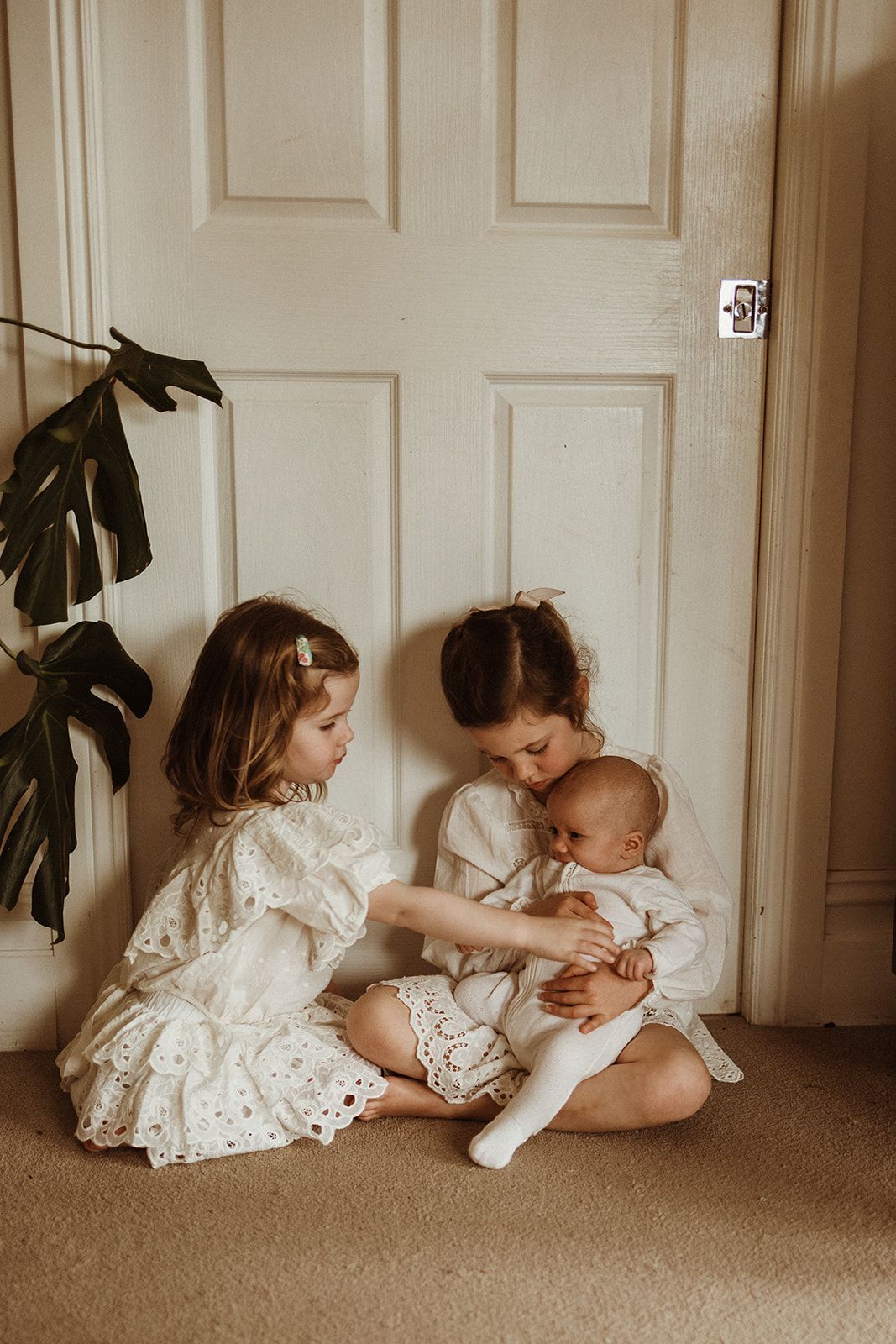 Two young girls in white dresses sit on floor cradling a newborn baby near a white door with a green plant nearby.