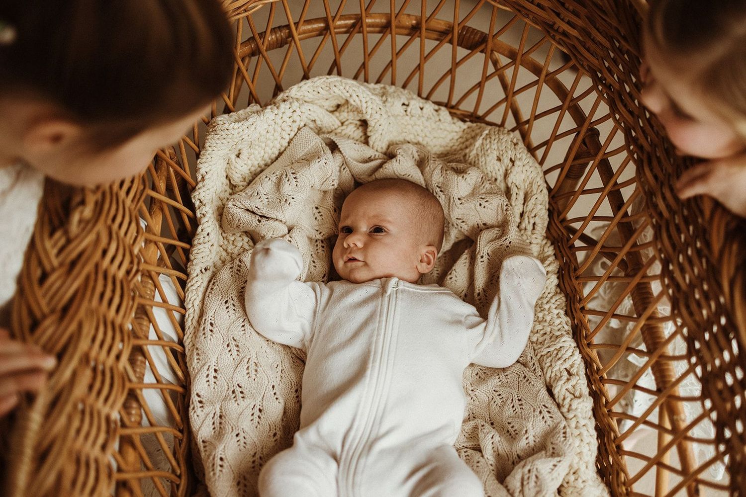 A peaceful newborn baby in a white onesie lying in a wicker bassinet with a cream knit blanket.