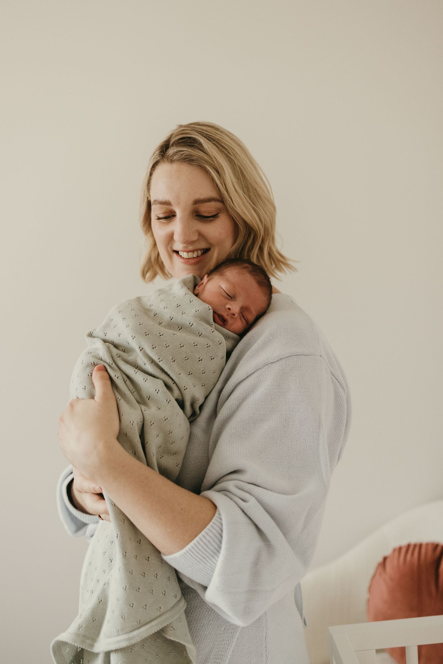 Intimate portrait of a parent sharing a joyful smile while holding a sleeping newborn wrapped in a soft gray blanket.