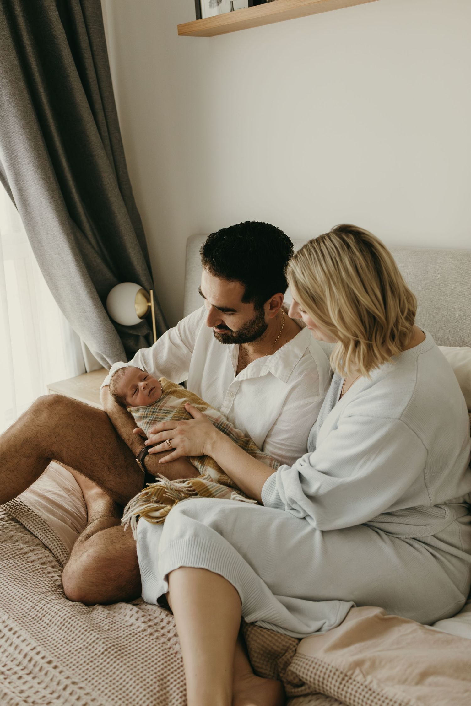 Couple snuggling with their newborn baby on a comfortable bed in natural light.