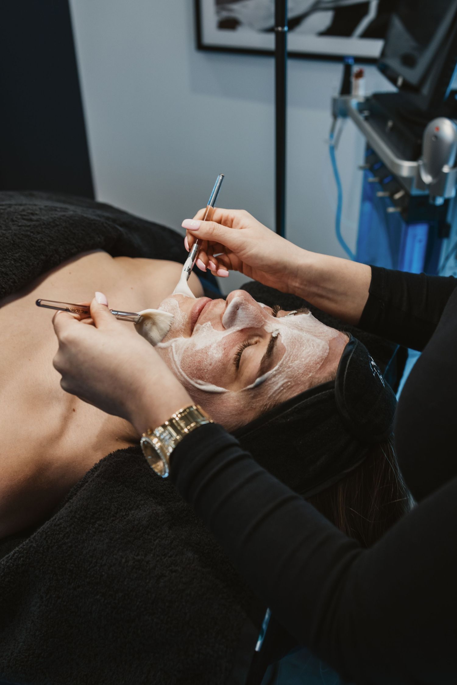 Esthetician applying white facial mask with brush to relaxed client lying on treatment bed in spa.