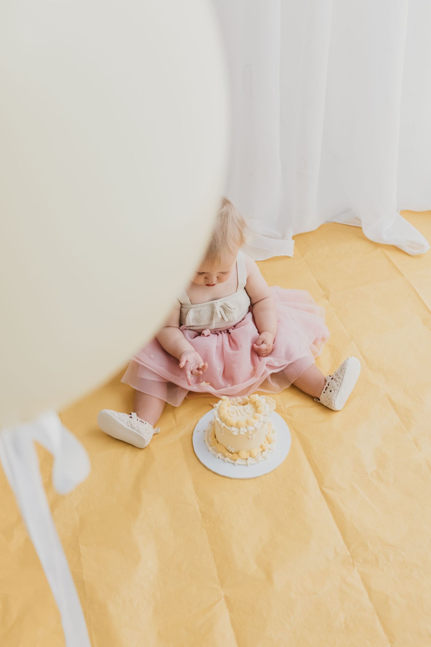 Baby girl in pink tutu dress sitting on yellow floor with smash cake during first birthday photo session.