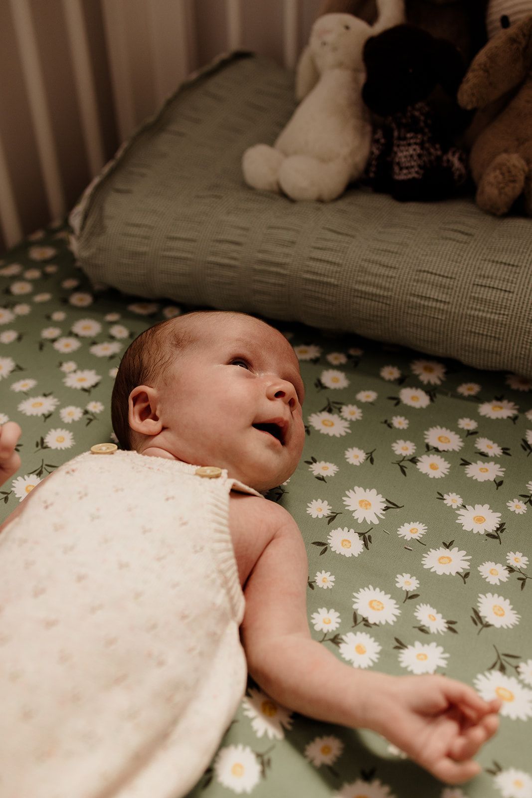 Newborn baby lying on a daisy-patterned green crib sheet, looking up, with stuffed animals in the background.