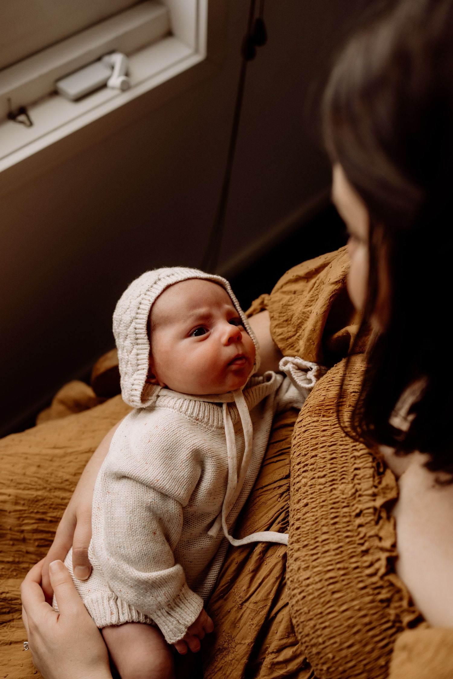 A newborn baby in a white knit romper and bonnet gazes up at their dark-haired mother in a warm, cozy setting.