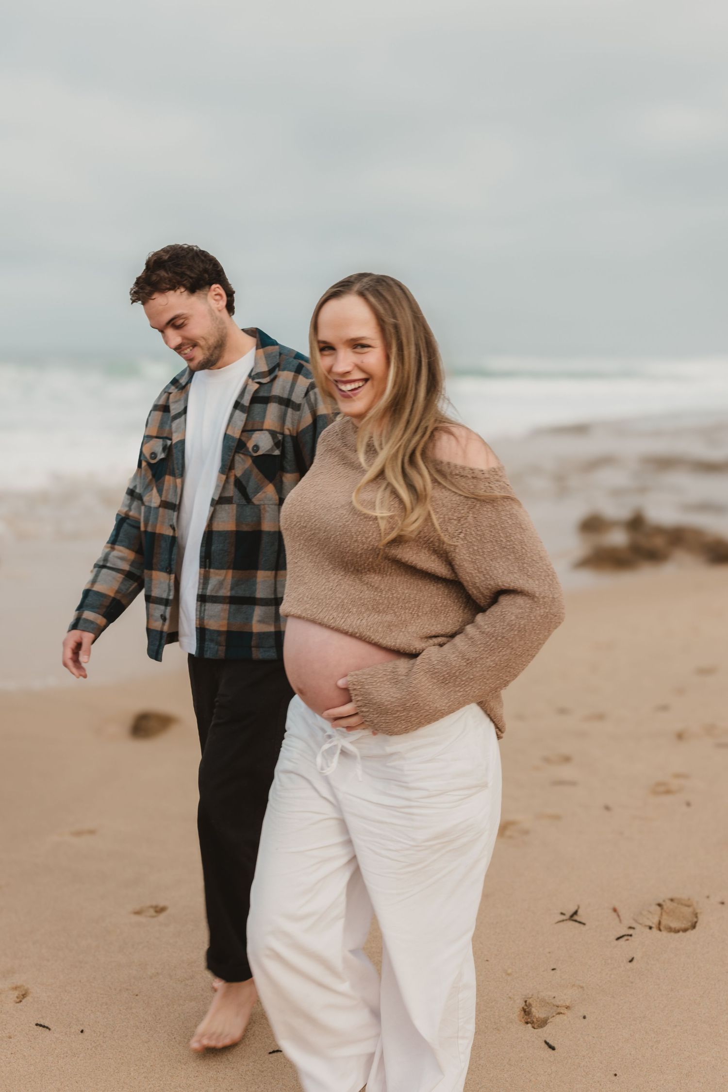 Pregnant woman smiling on beach with partner, wearing beige sweater and white pants, ocean waves in background.