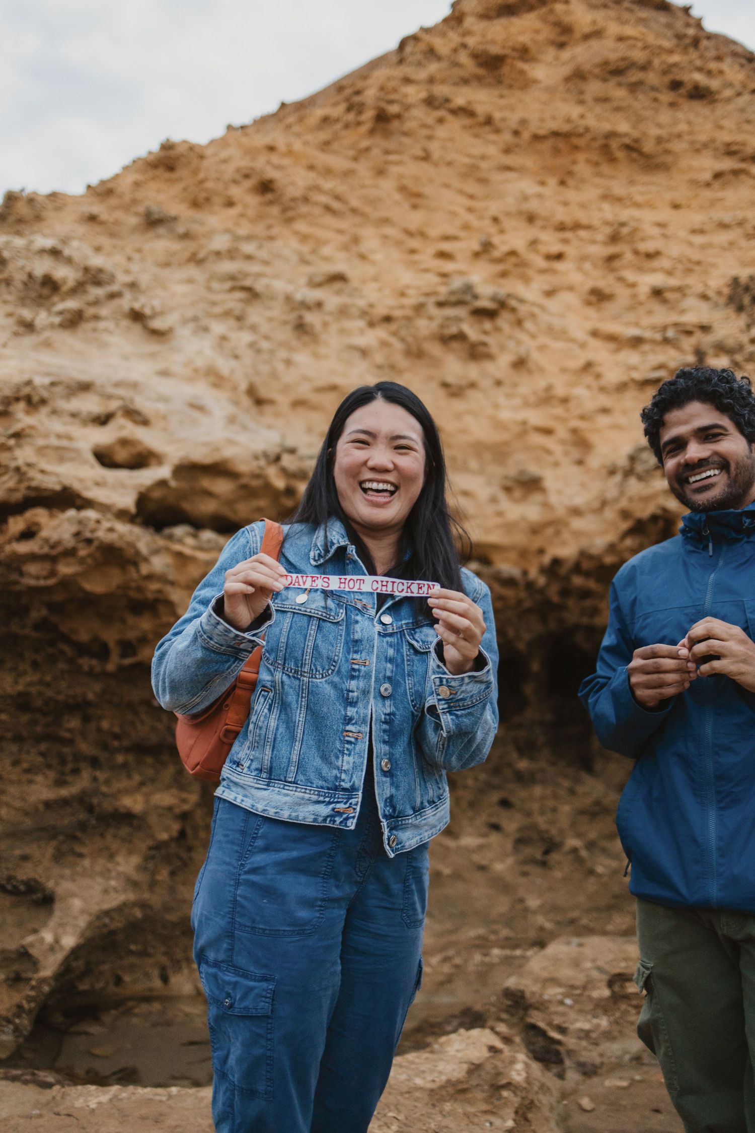Two happy friends laughing outdoors near rocky cliffs, woman in denim jacket holding a bracelet.