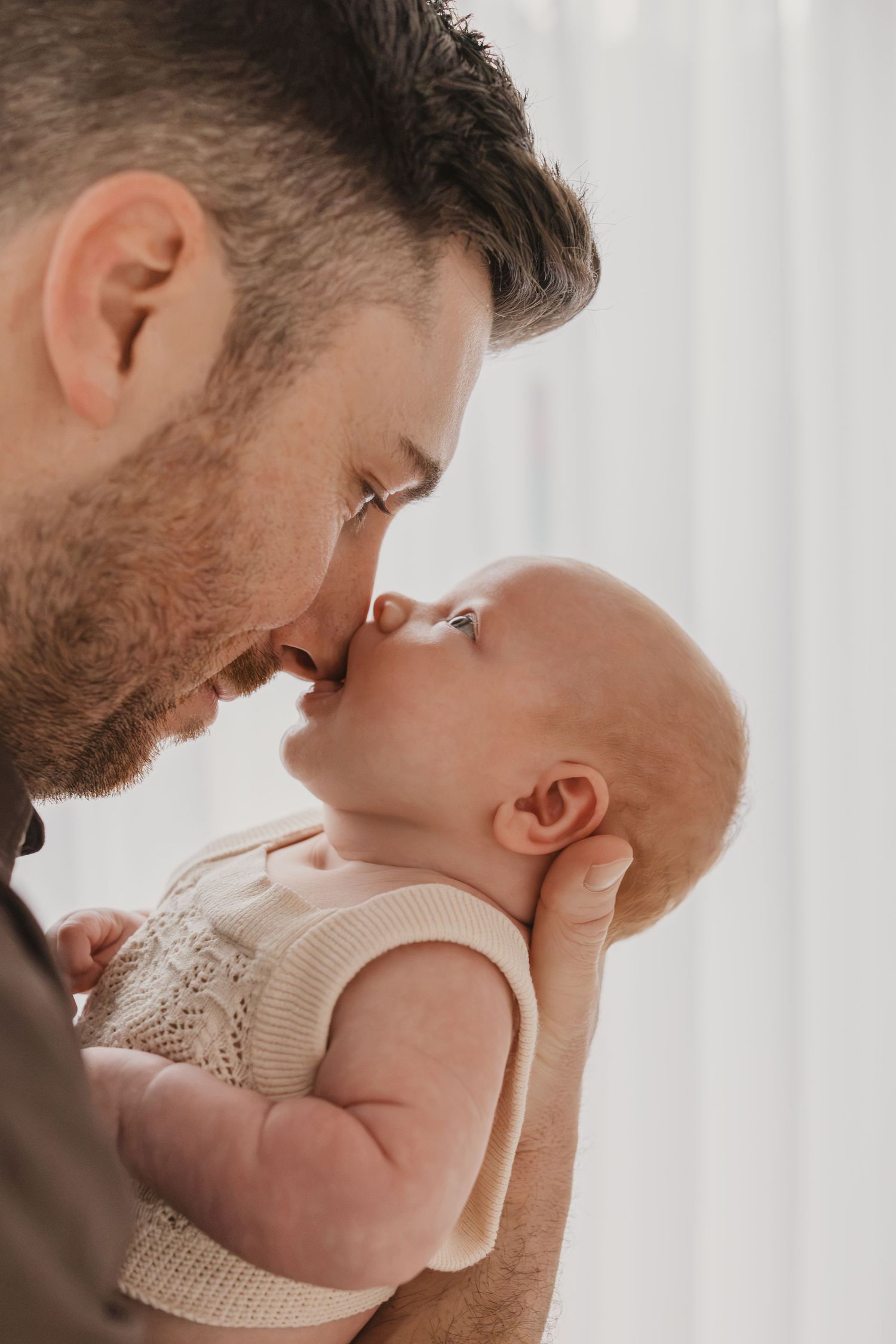 A bearded father tenderly touches noses with his newborn baby, sharing a sweet bonding moment.