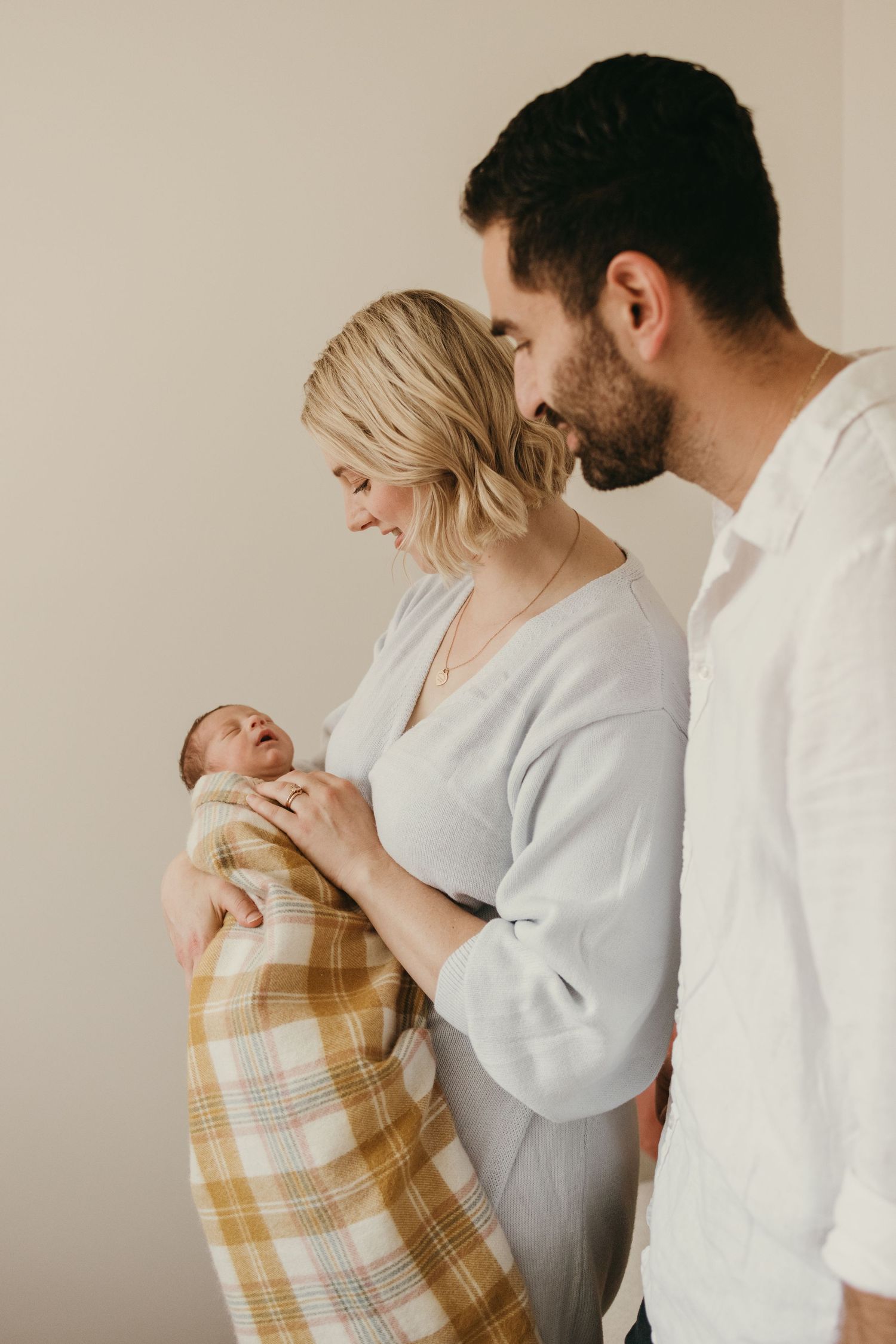 Intimate family moment with newborn wrapped in plaid blanket against neutral wall.