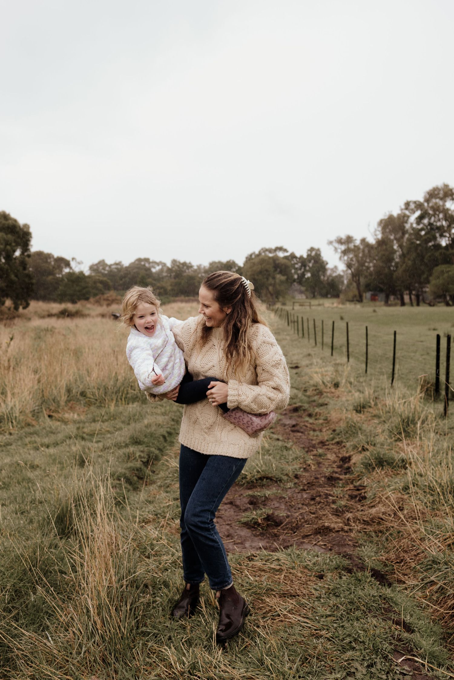 A smiling woman in a beige sweater holds a toddler while walking along a rural fence path in a grassy field.
