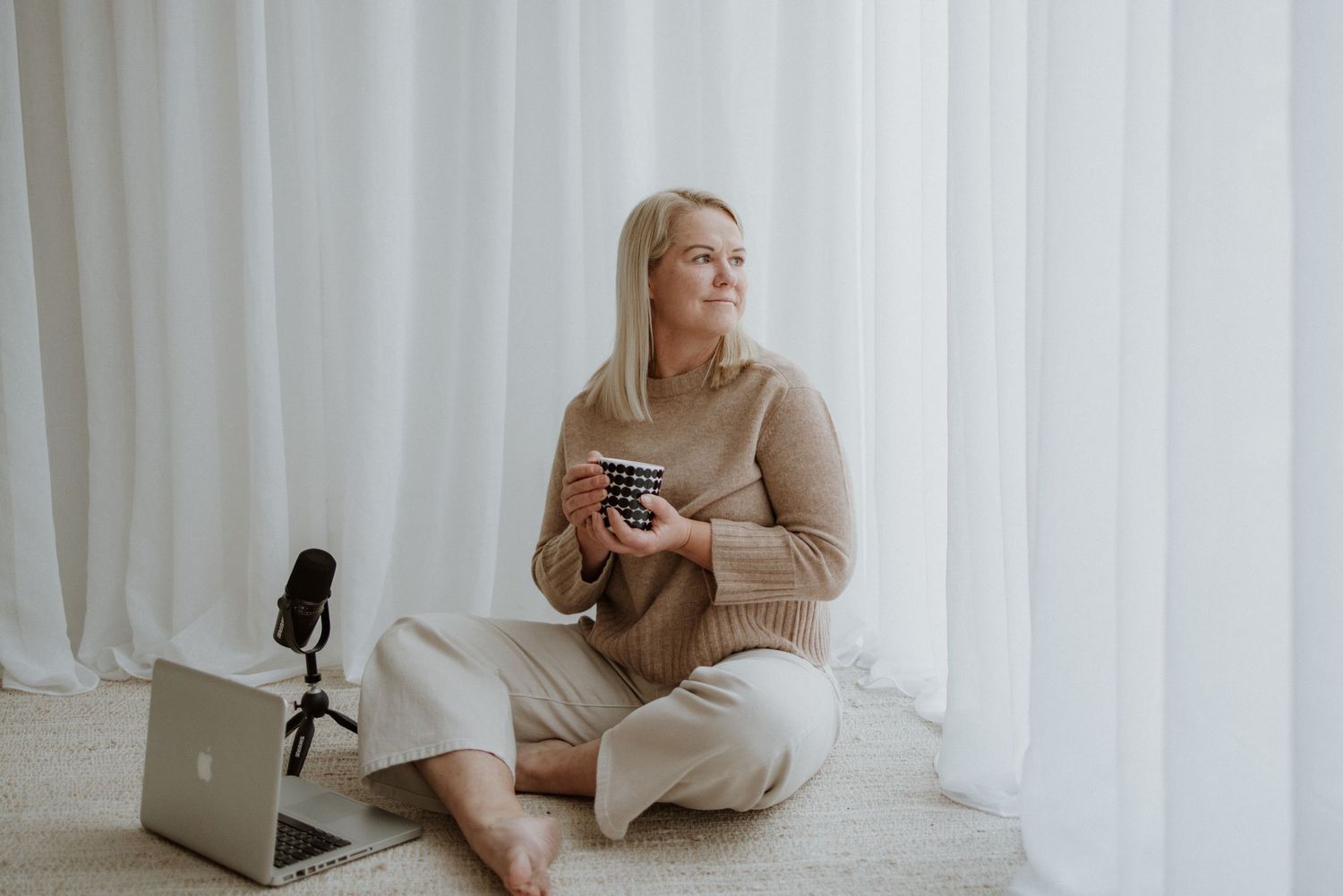 Woman in beige sweater sitting cross-legged on floor holding coffee cup near laptop and microphone by white curtains.