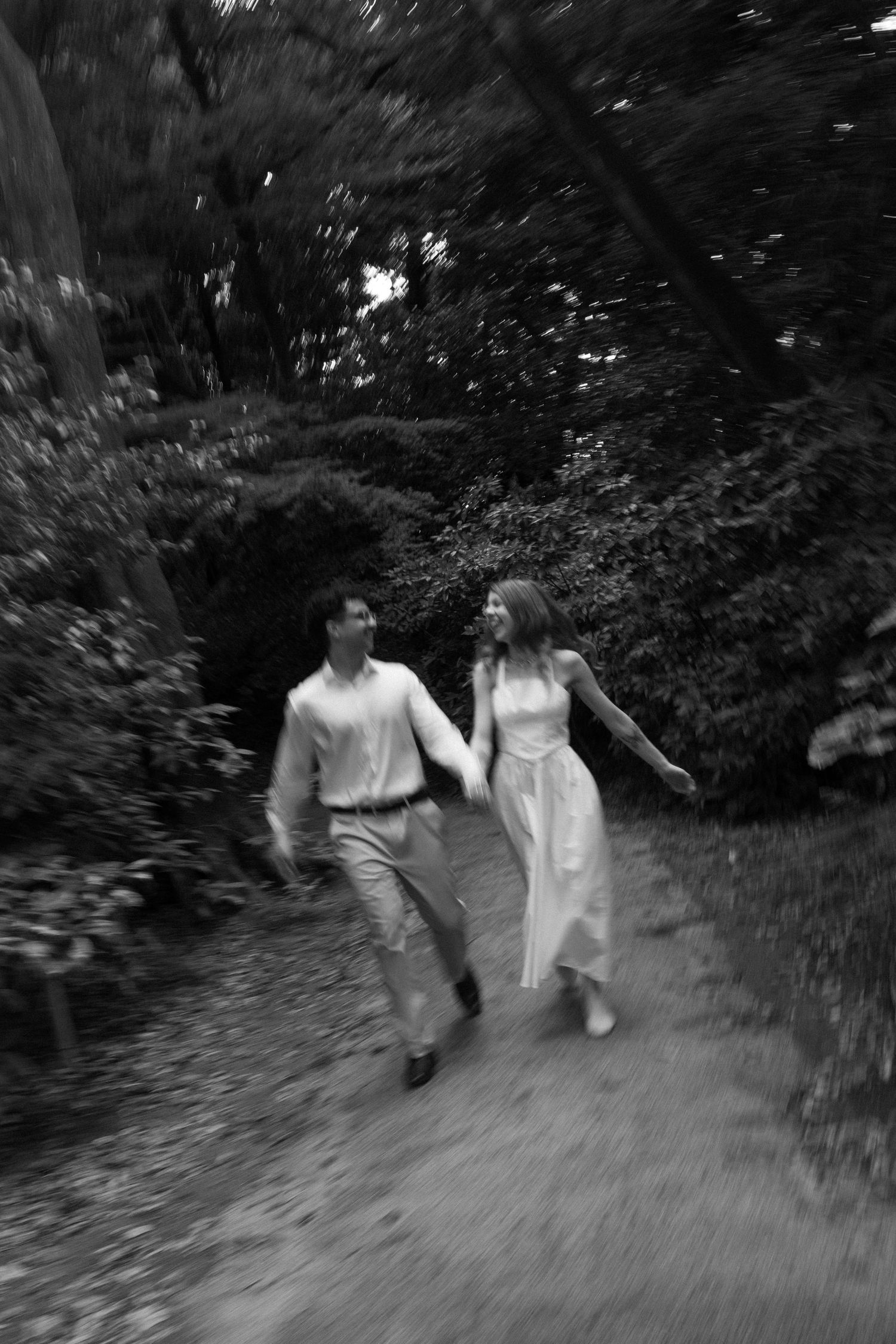 Black and white photo of a couple holding hands and running joyfully along a tree-lined garden path.