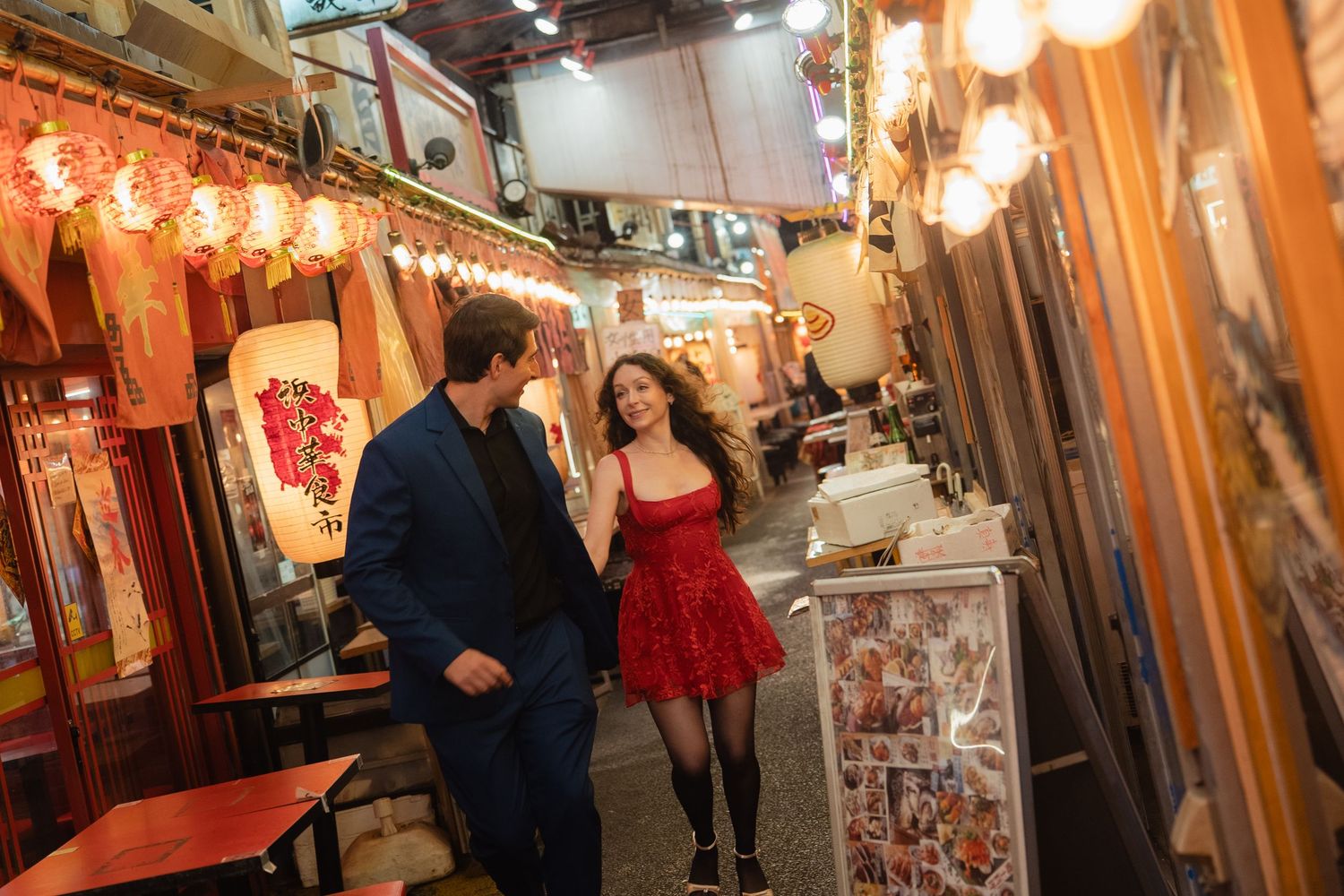 Couple walking hand in hand through a warmly lit Japanese alley with lanterns and restaurant signs at night.