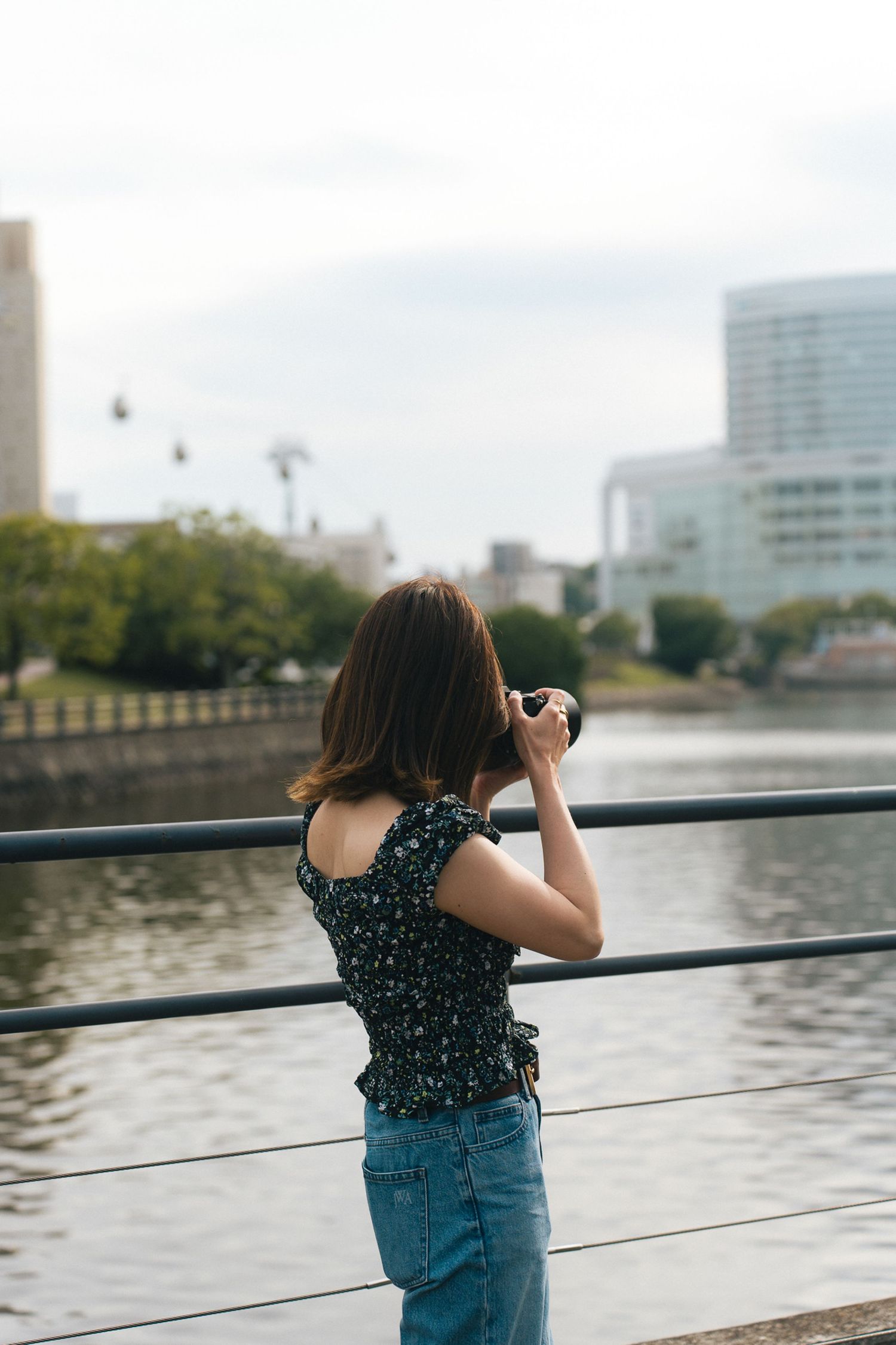 Young woman in floral top photographing city skyline and river from a waterfront railing.