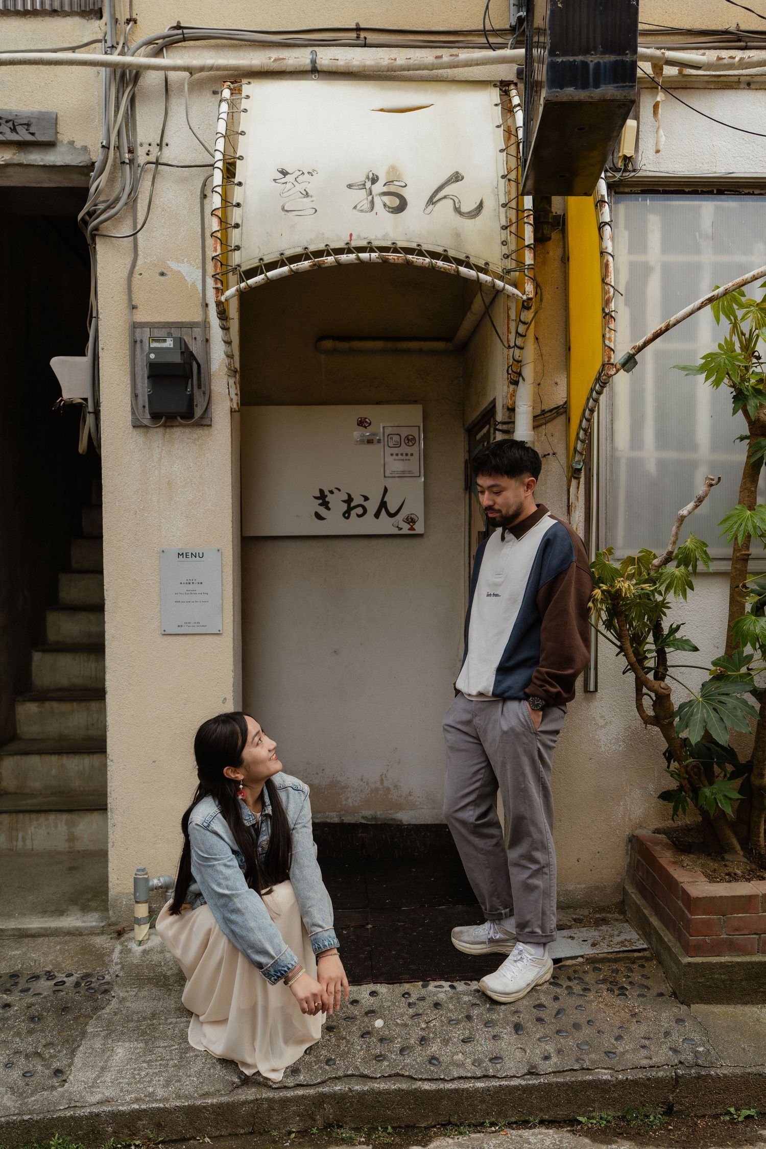 Couple posing outside a traditional Japanese building with kanji signage in an urban alleyway setting.