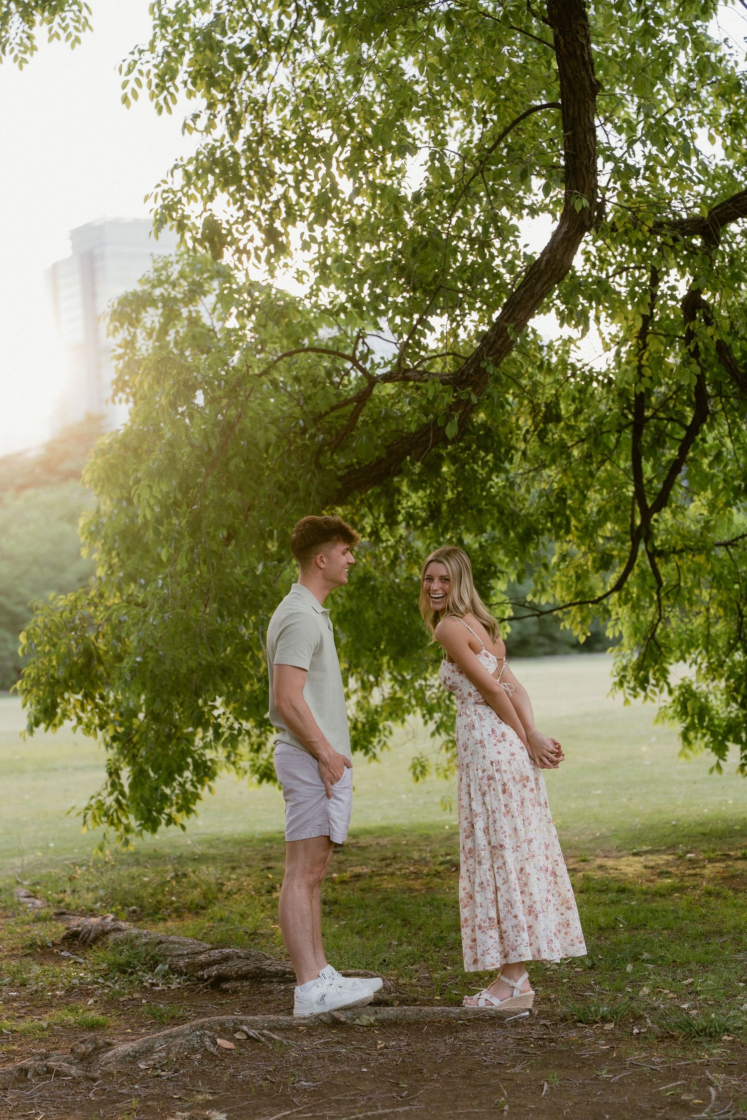 Couple standing under a green tree in a sunny park, woman in floral dress smiling at man in casual summer outfit.