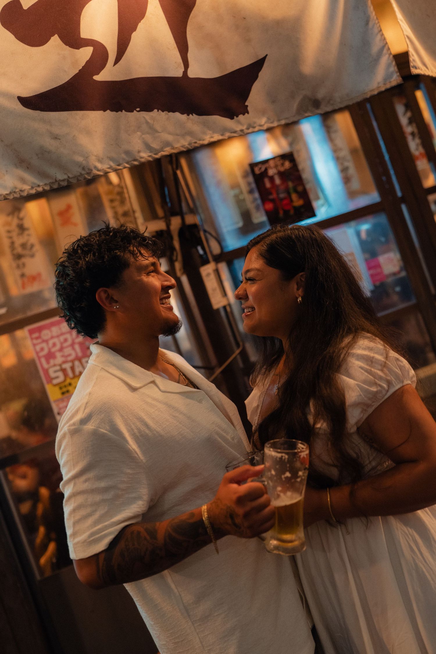 Couple smiling at each other holding beer glasses outside a Japanese restaurant at night.