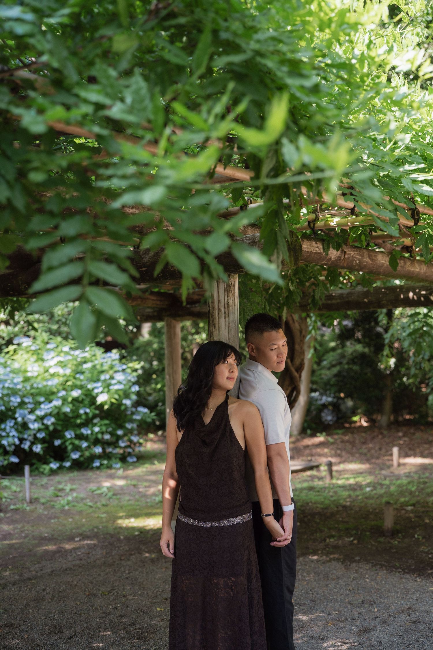 Couple standing back-to-back under a wooden pergola with lush green vines in a serene garden setting.