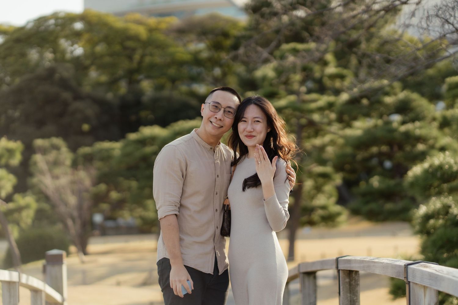 A smiling couple poses together on a wooden bridge surrounded by lush green trees in a scenic outdoor setting.