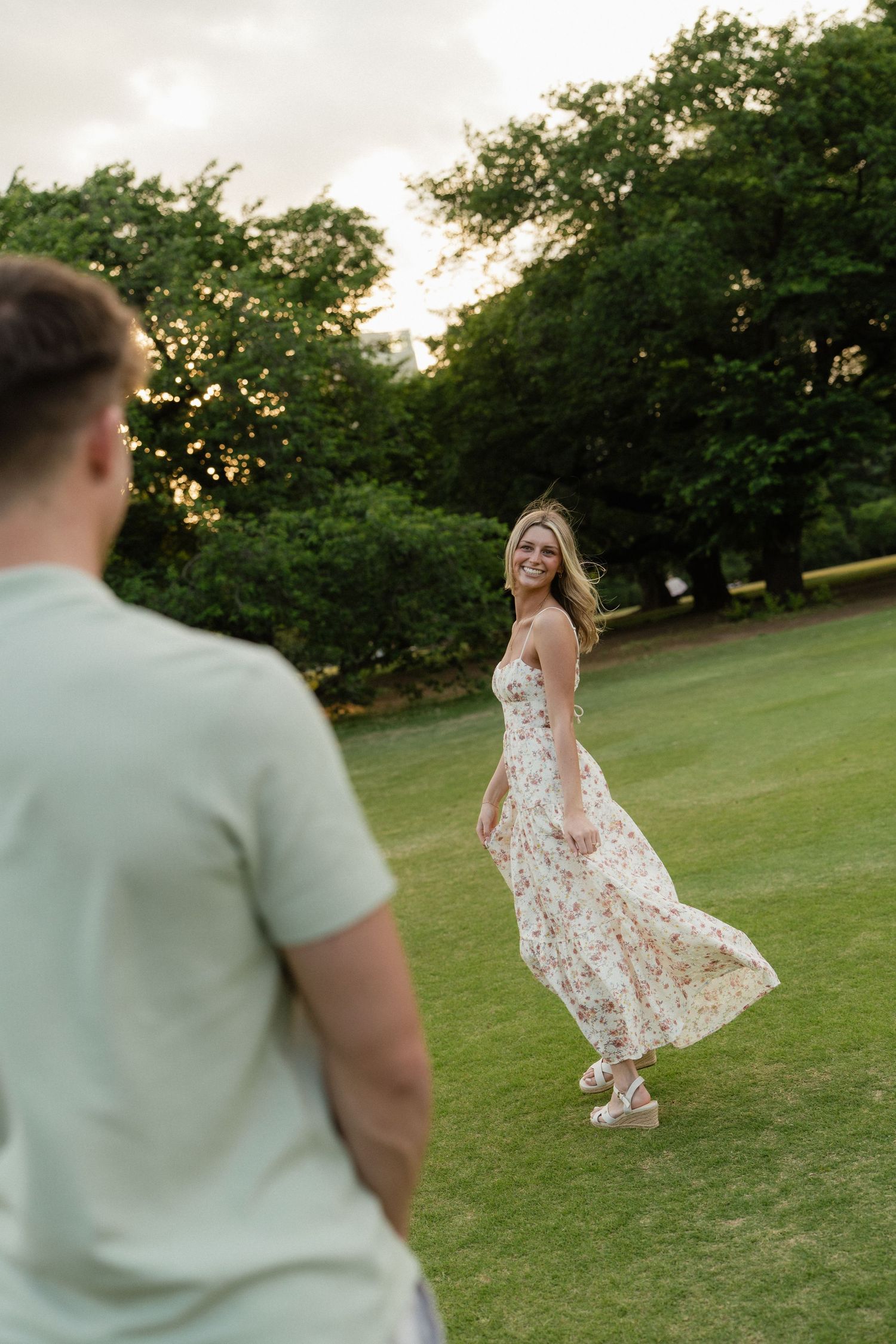 A smiling woman in a floral dress twirls on a green lawn while a man in a light shirt watches her.