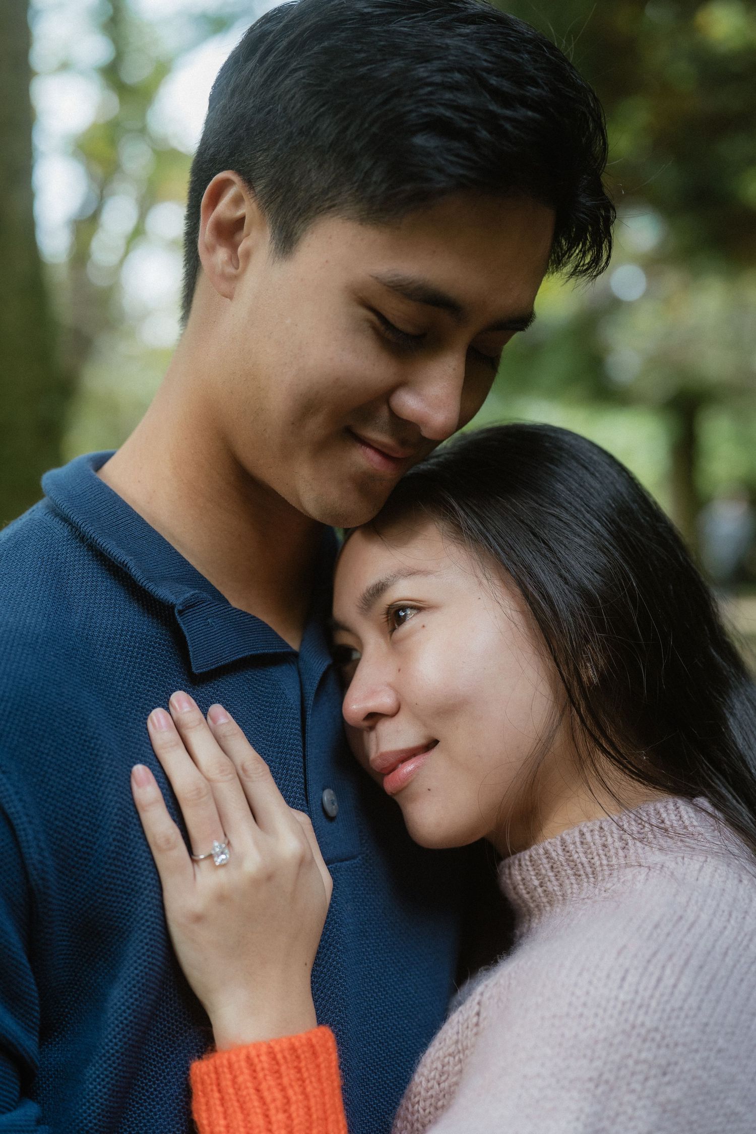 A loving Asian couple embraces tenderly outdoors, the woman resting her head on the mans chest while showing an engagement ring.